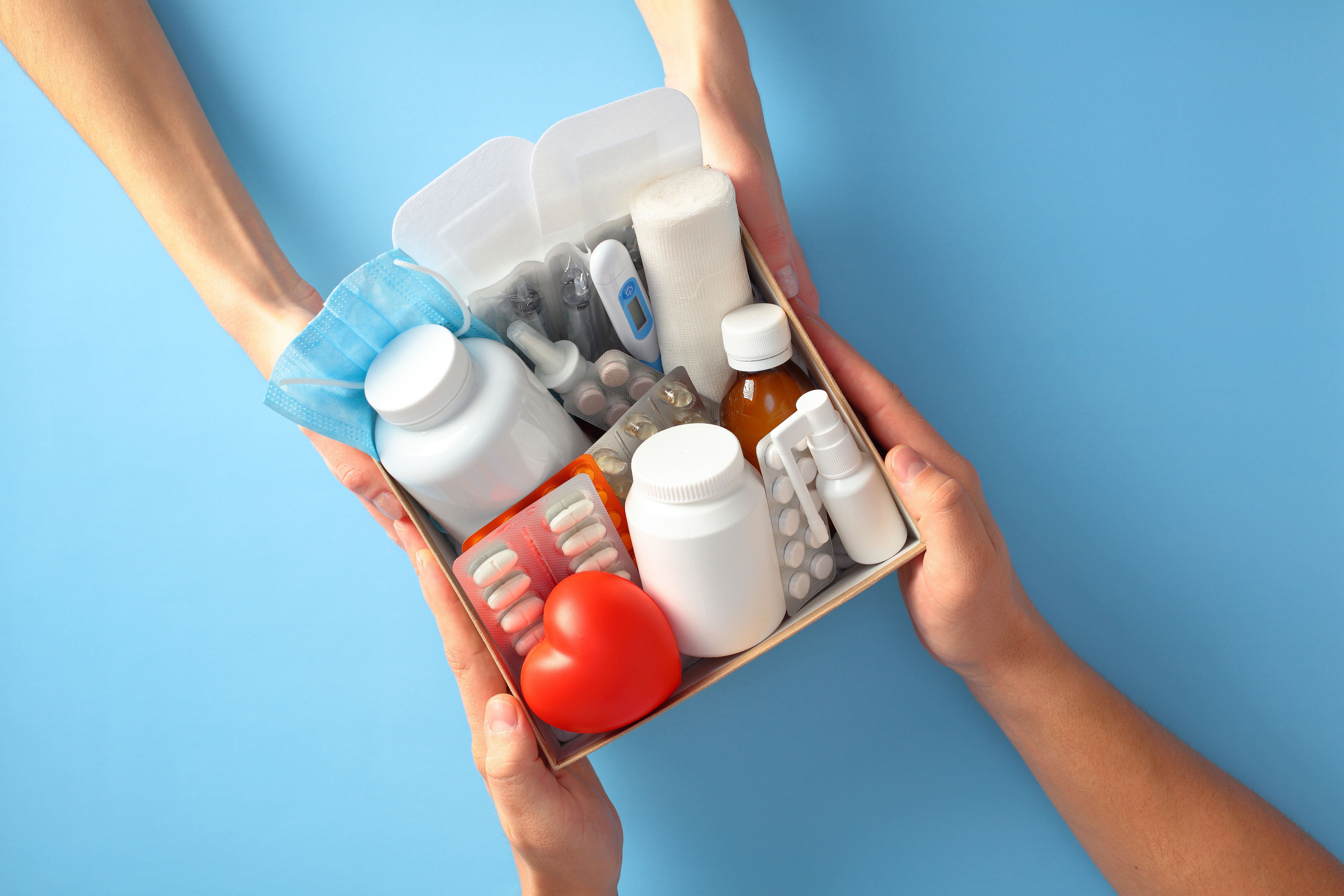 Hands holding a healthcare kit with medication, pills, thermometer, and bandages on a blue background. Home medication delivery, online pharmacy, donations of medicines, first aid box concept.