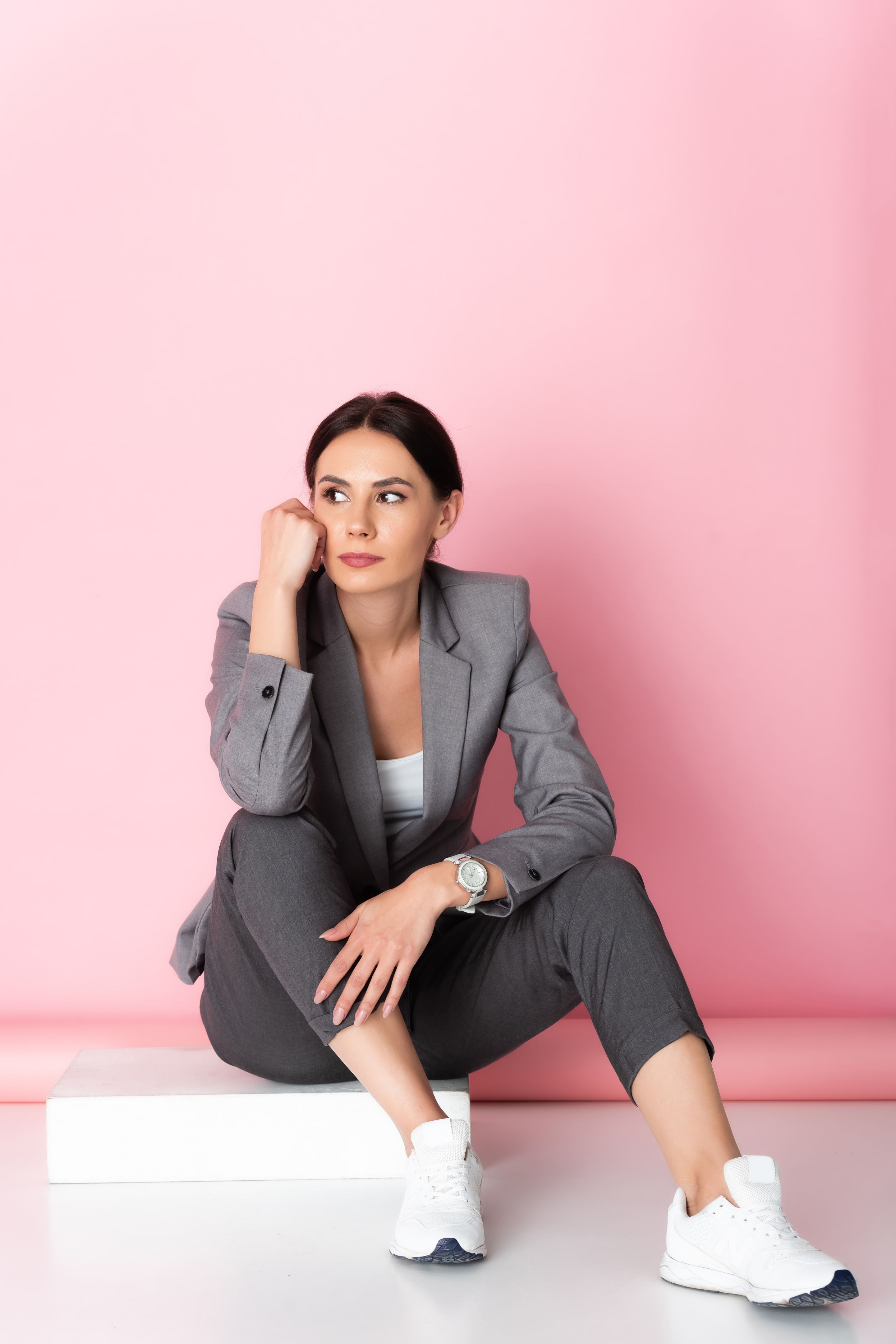 pensive businesswoman in suit and white sneakers looking away while sitting on pink