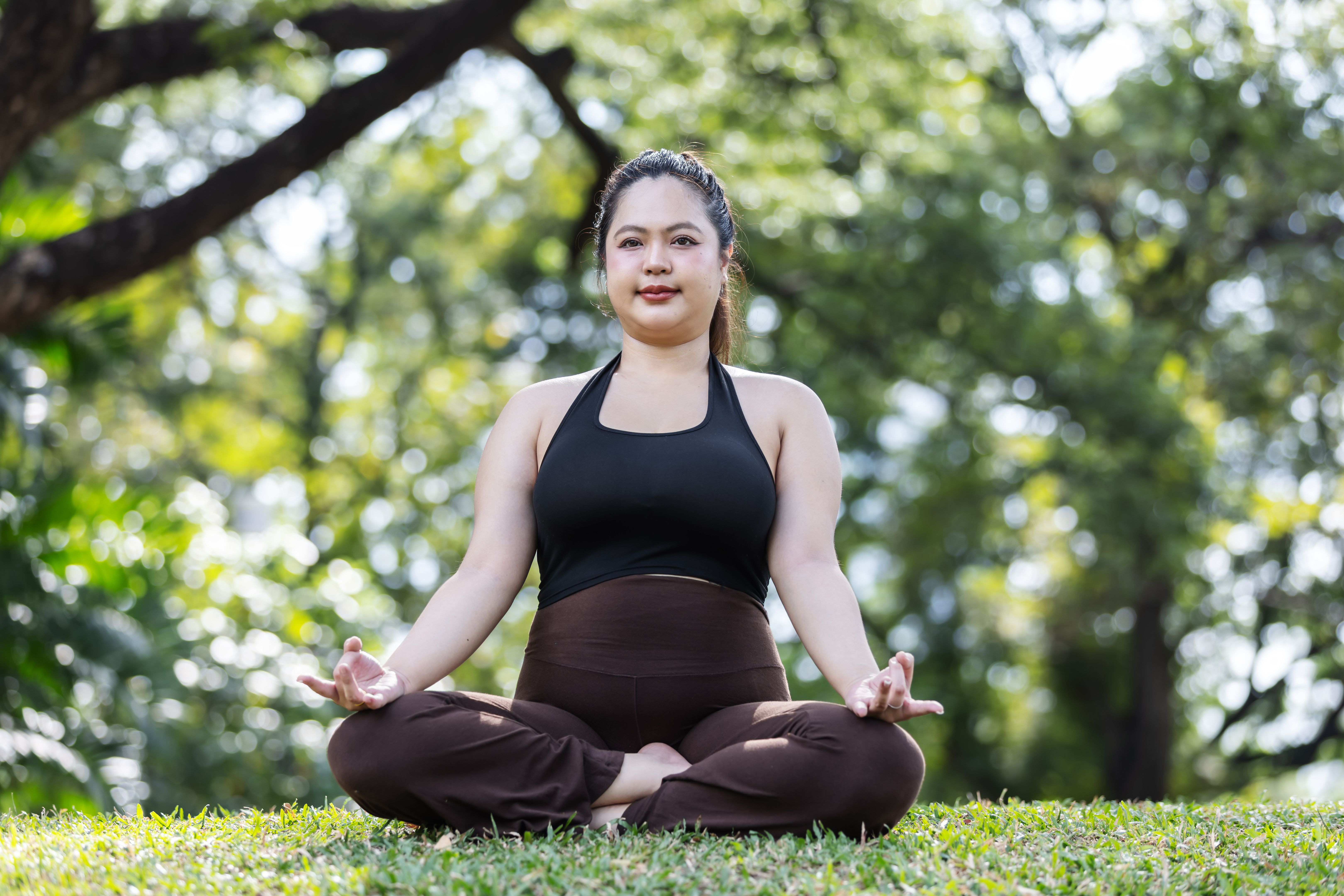 A calm plus-size Asian woman sits cross-legged in meditation on green grass, practicing mindfulness and balance. A peaceful outdoor wellness scene that promotes body positivity, mental health, and inner strength.