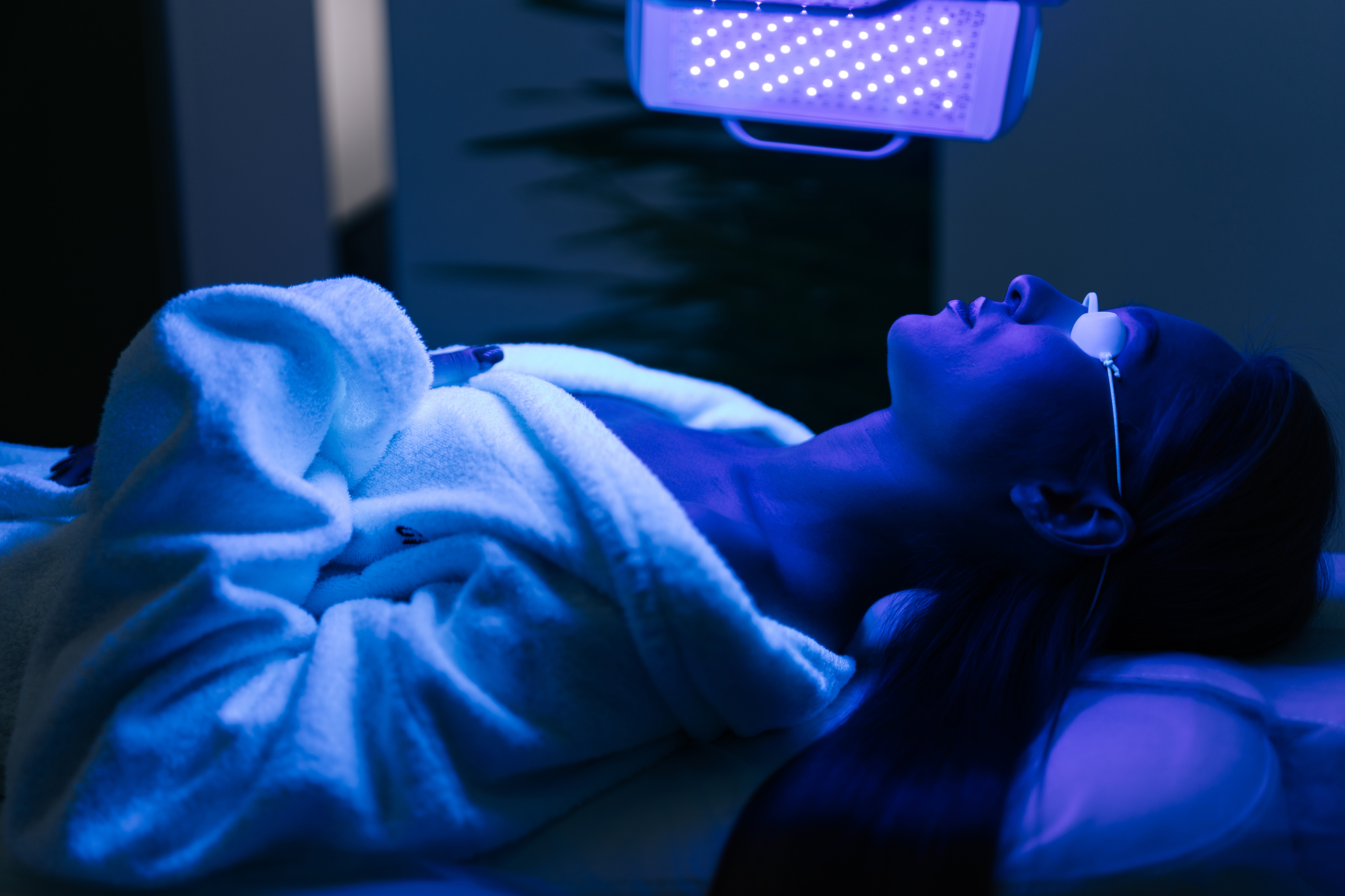 Young woman in glasses having blue LED light facial photodynamic therapy treatment in beauty salon. Female client having non-invasive type of phototherapy. Young woman in glasses having blue LED light facial photodynamic therapy treatment in beauty salon. Female client having non-invasive type of phototherapy.