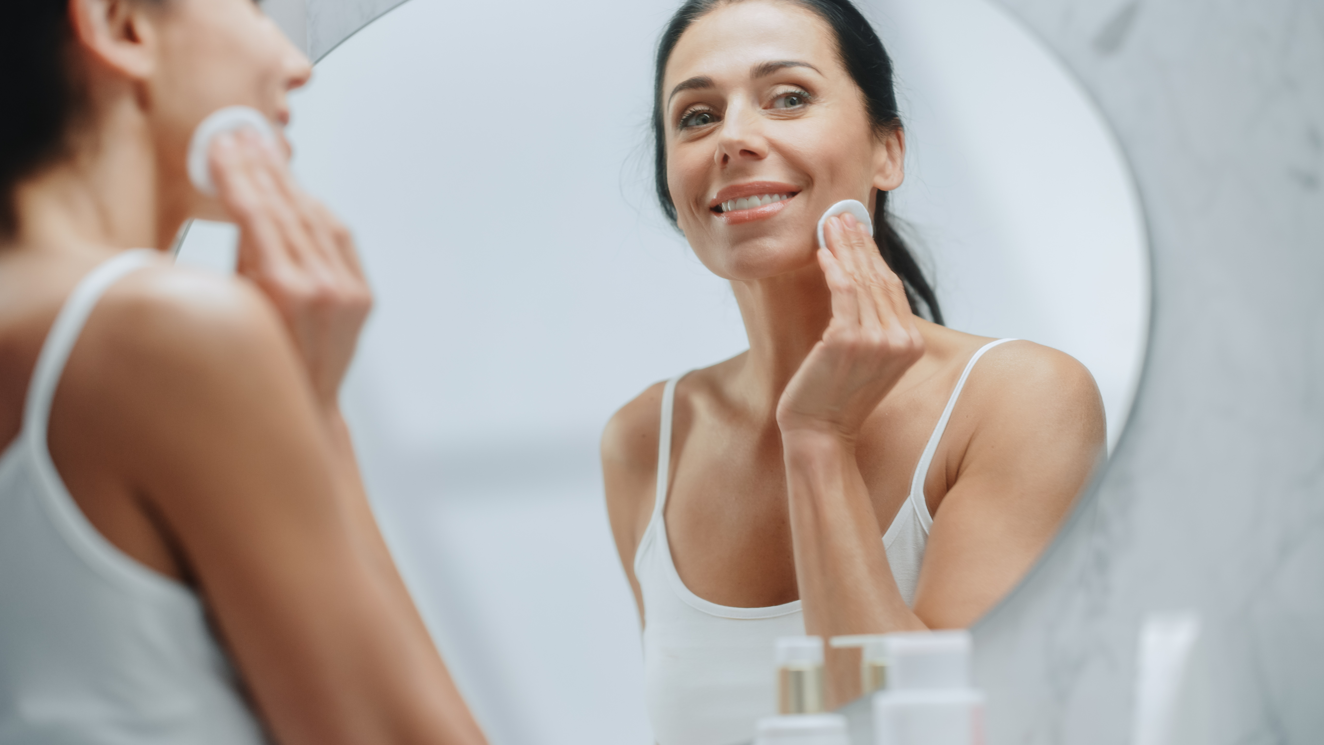Beautiful Caucasian Woman Uses Cotton Pad to Clean Face Makeup, Skin, Smiles in the Mirror. Happy Middle Aged Female Enjoying Her Beauty Routine. Wellness, Natural Beauty, Cosmetic Skincare Products