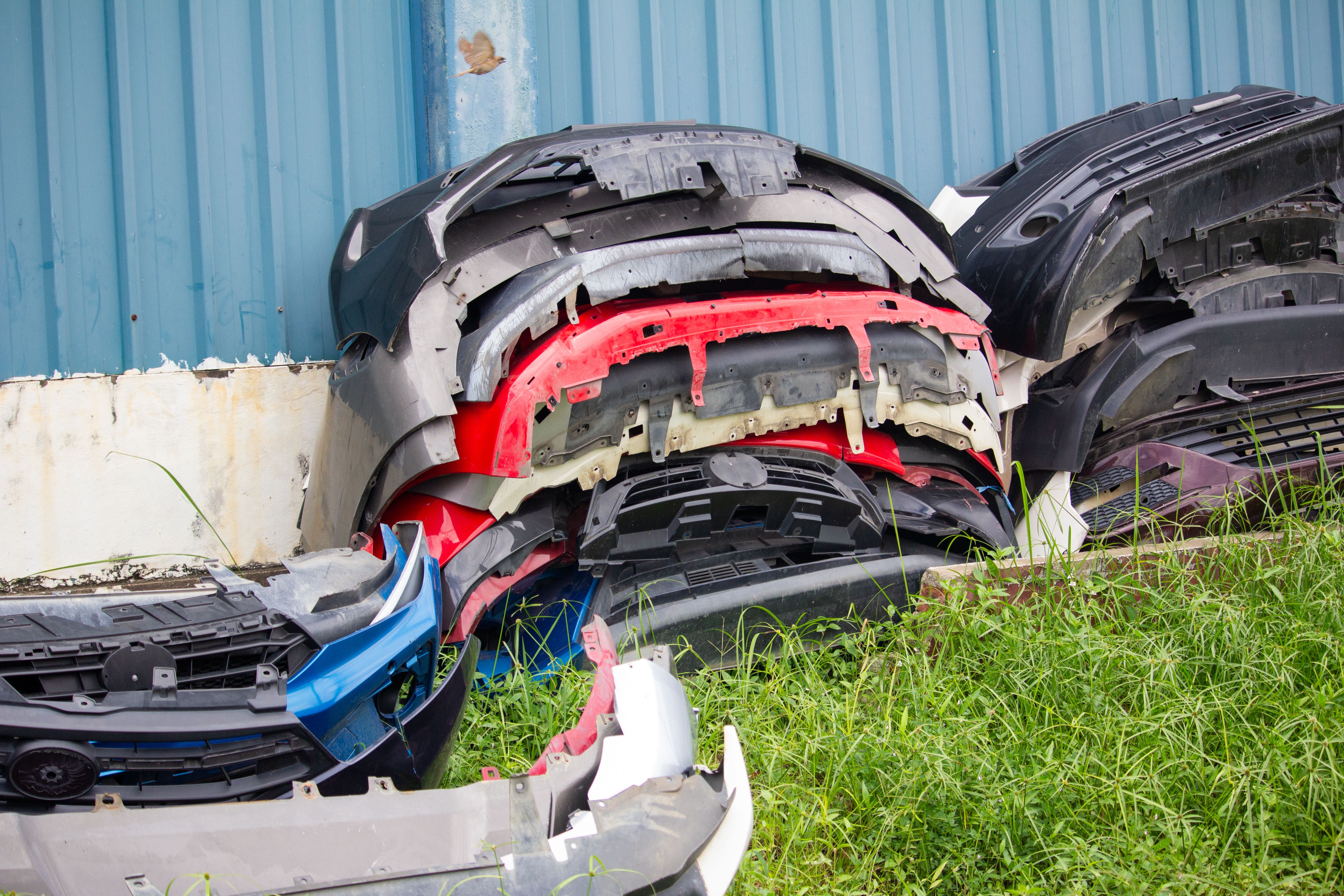 A pile of colorful used car bumpers stacked near a wall on grassy ground at an auto repair or recycling yard outdoors