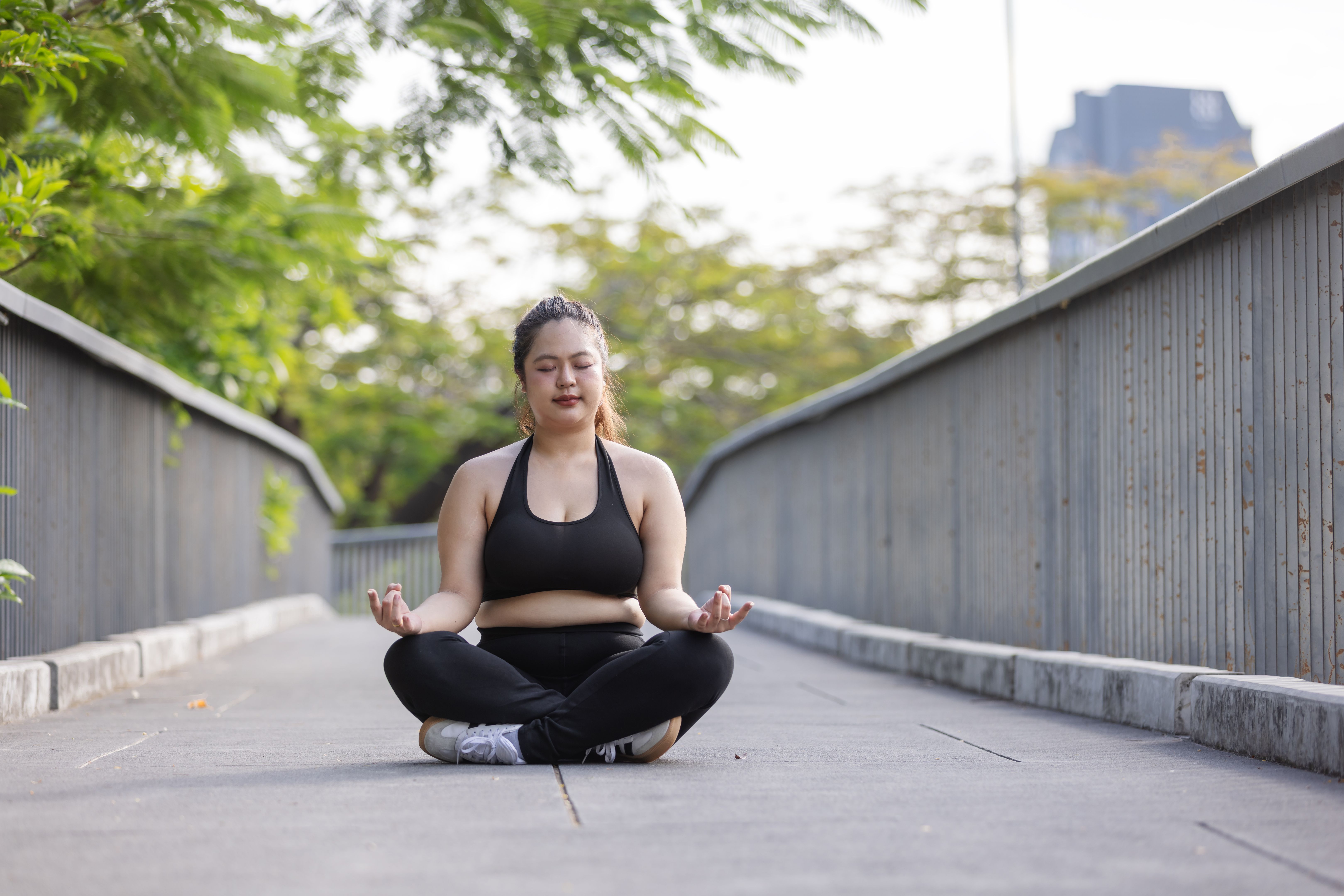 A calm plus-size Asian woman sits cross-legged in meditation on an outdoor urban walkway, practicing mindfulness and balance. A modern wellness scene promoting body positivity, mental health, and peaceful active living.