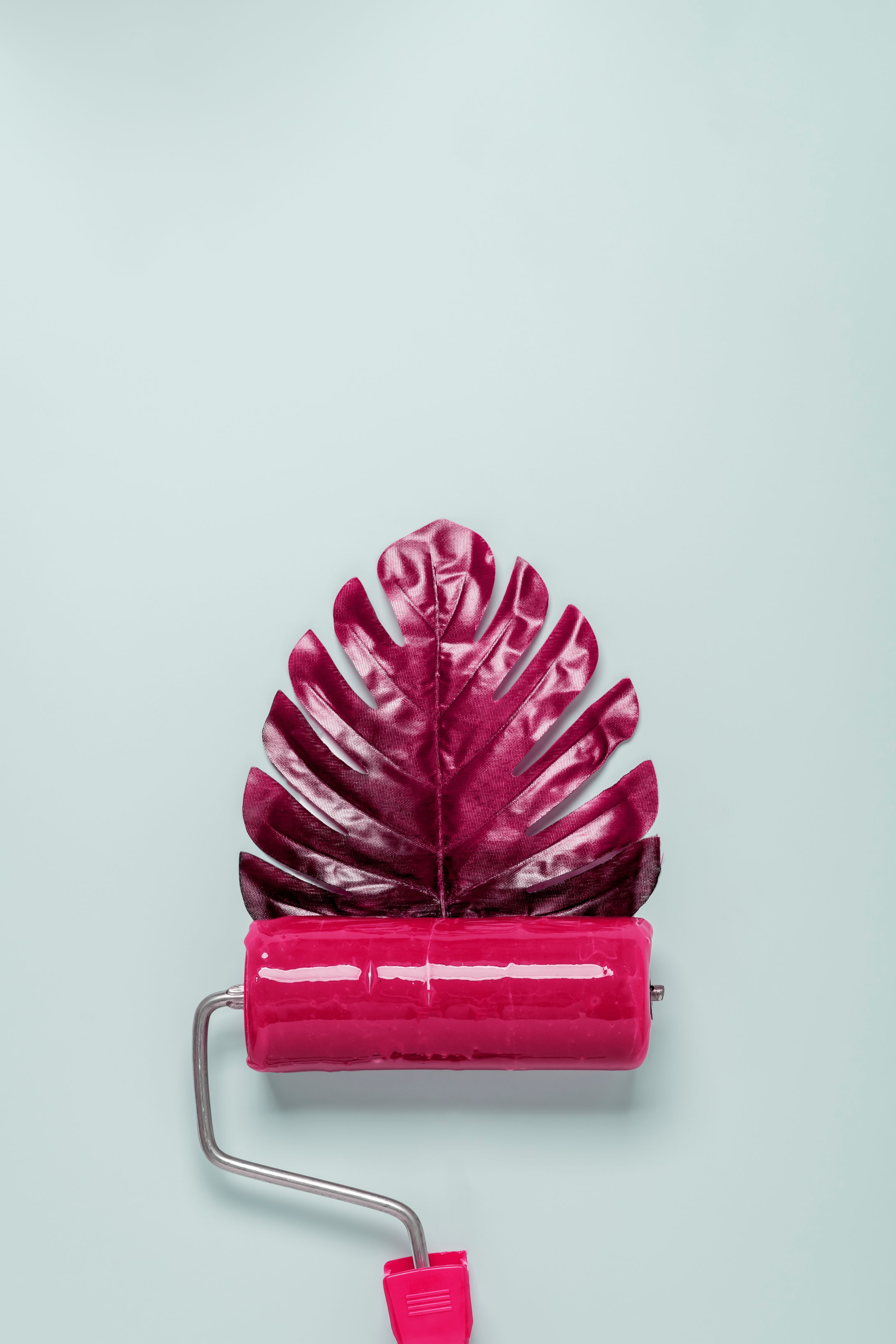 A paint roller with red paint creates a red monstera leaf on a pastel blue background.