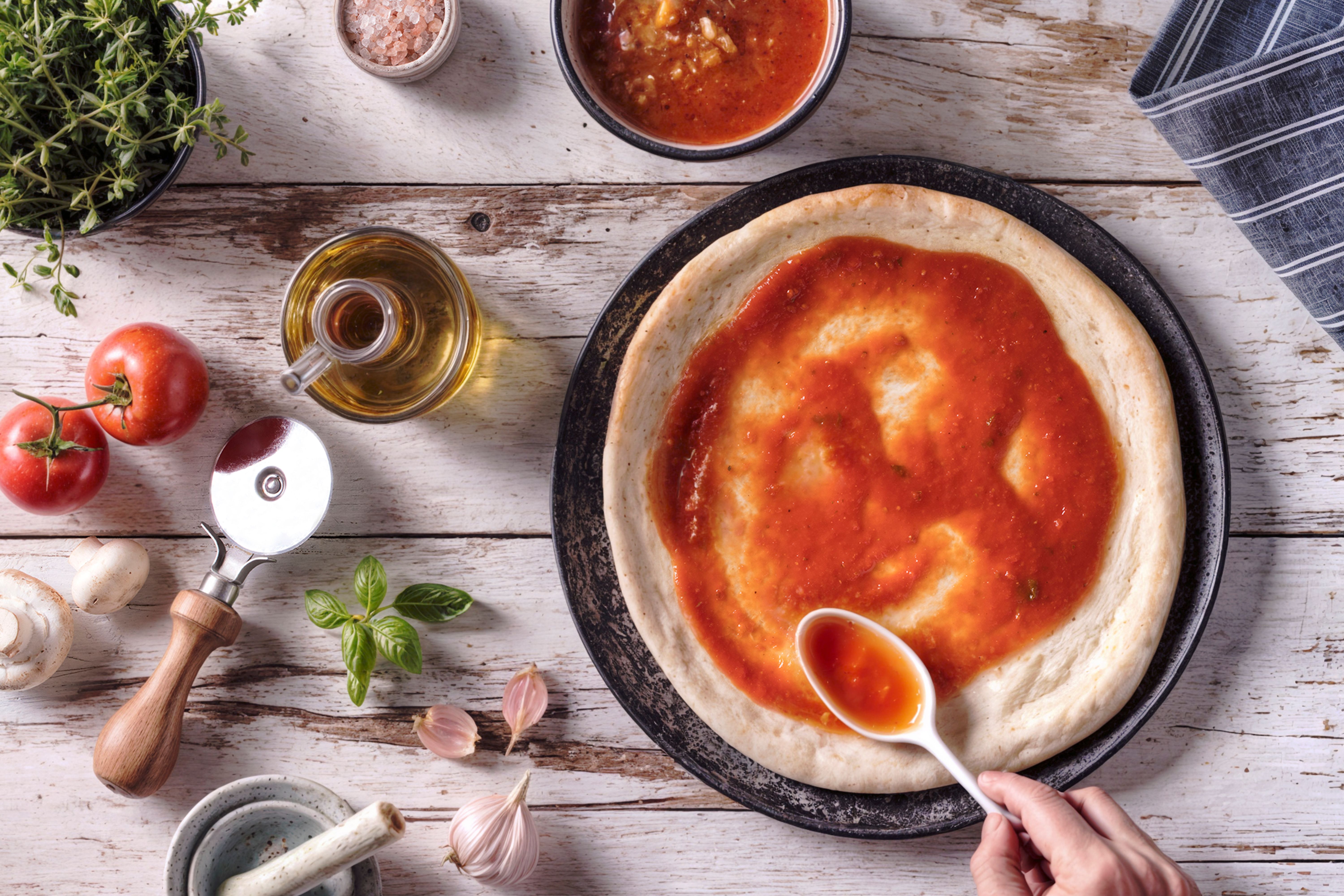 A vibrant scene of pizza preparation featuring fresh tomatoes, herbs, olive oil, and a hand spreading rich tomato sauce on dough, perfect for Italian cuisine lovers.
