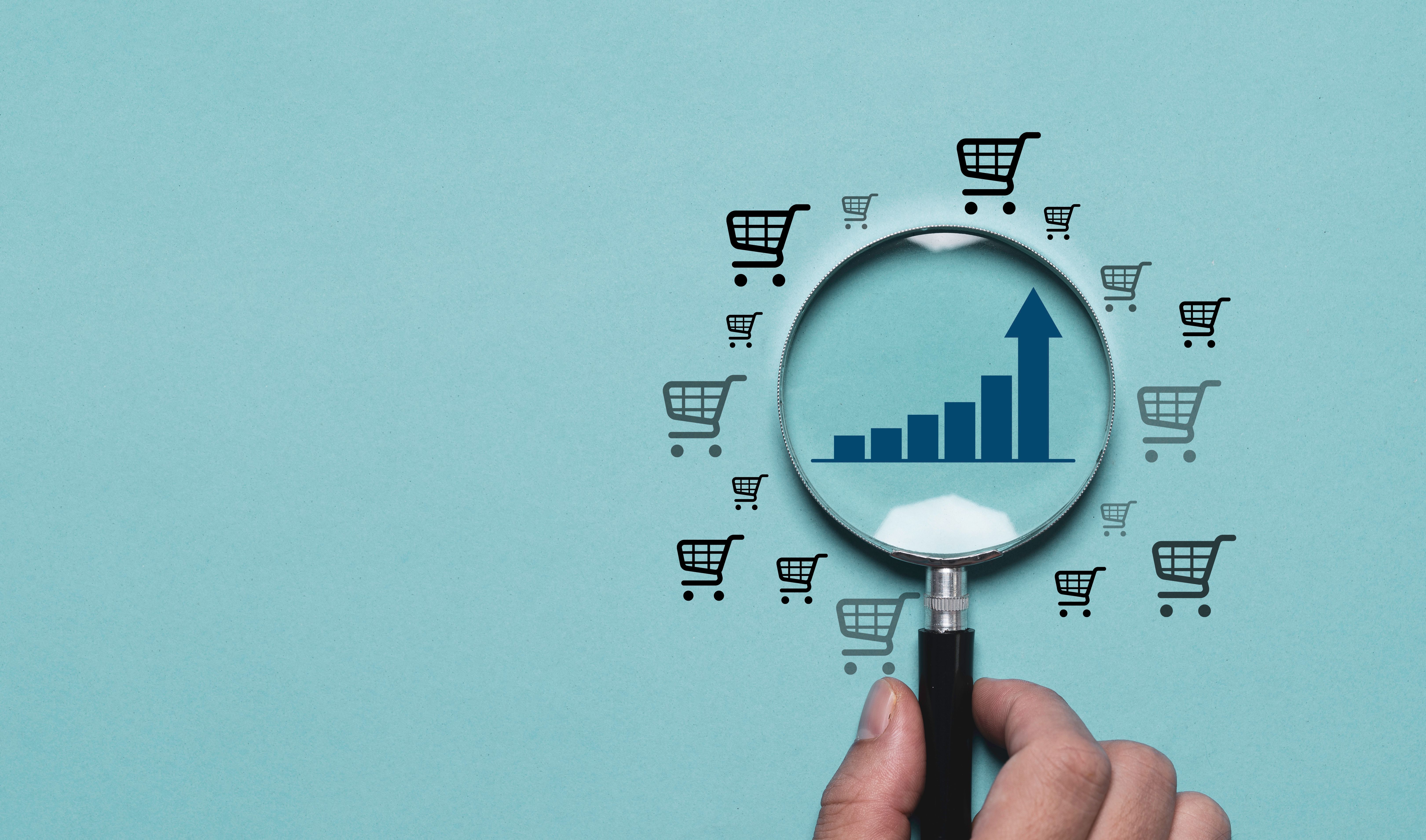 A hand holds a magnifying glass focusing on a blue bar graph with an upward trend, surrounded by cart icons on a light blue background. This symbolizes e-commerce growth and market analysis concept.