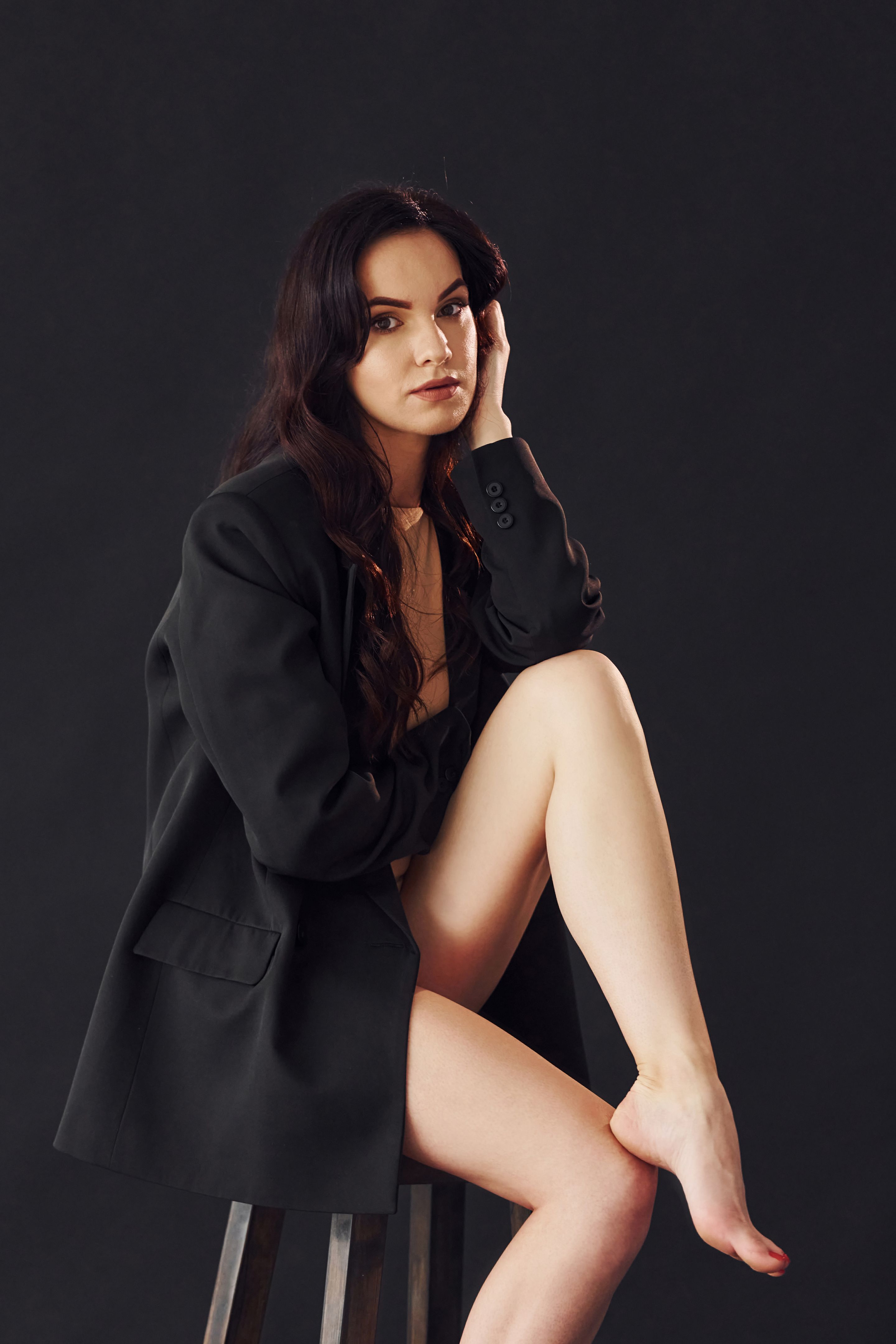 Sexy brunette with curly hair in luxury clothes sits on chair in the studio against black background