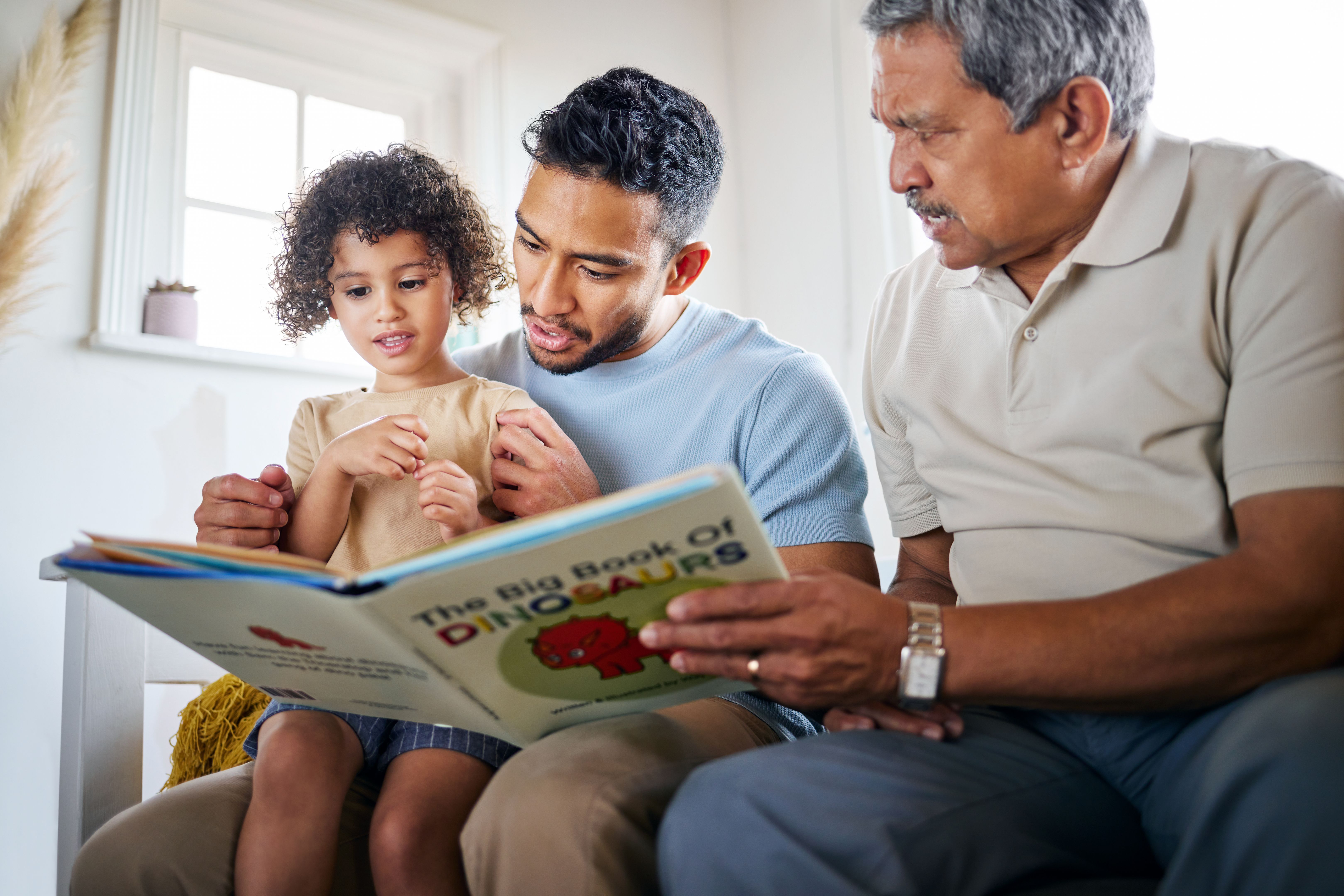 Shot of a little girl reading a book with her father and grandfather at home Shot of a little girl reading a book with her father and grandfather at home
