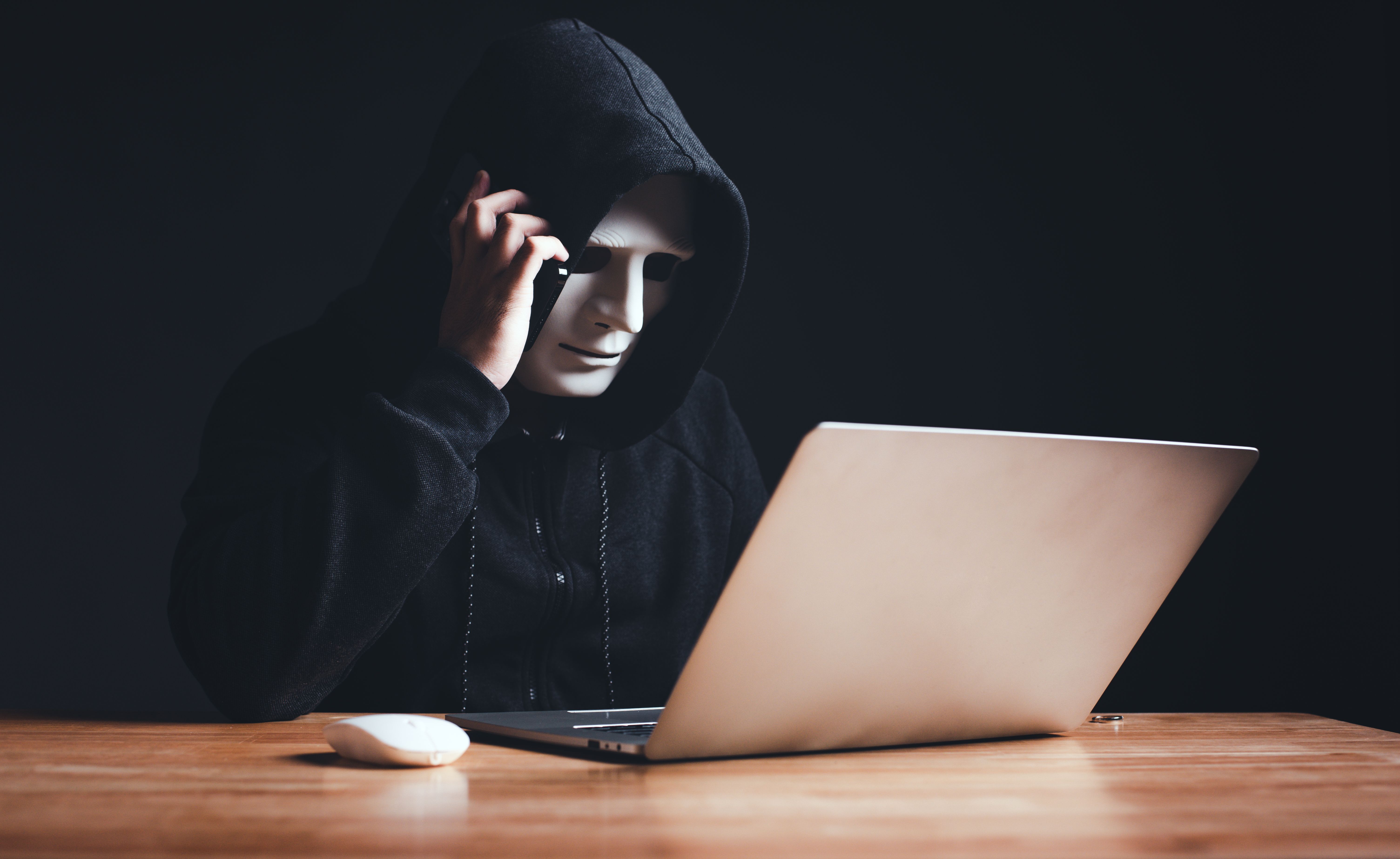 Black hat hacker in hood using laptop computer and call smartphone to victim on desk hacking privacy sensitive data hack in dark room background. Cyber security cyber crime concept. Hacking phishing