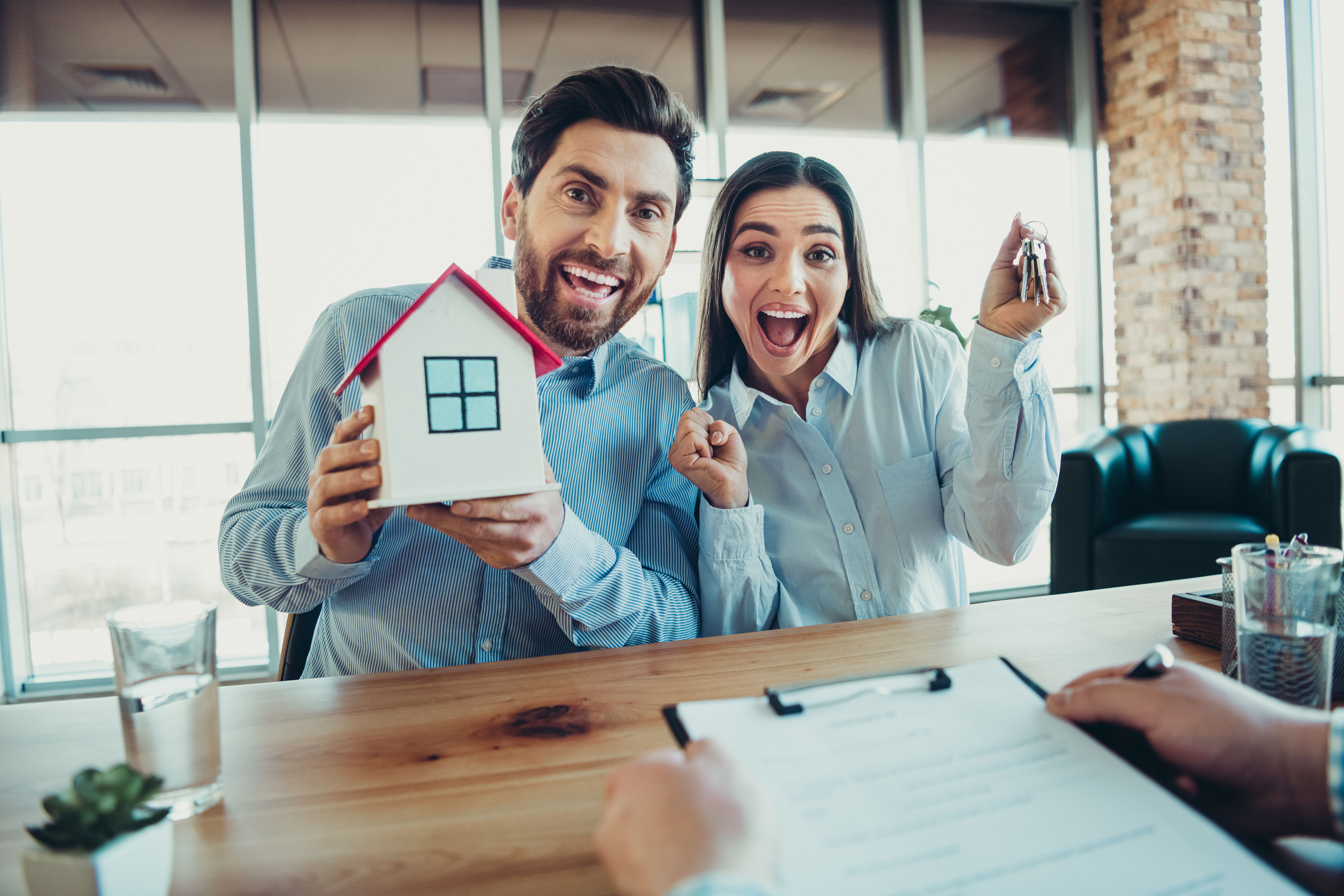 Excited young couple celebrates their new home purchase, showing house keys and a model, inside a bright workspace Excited young couple celebrates their new home purchase, showing house keys and a model, inside a bright workspace