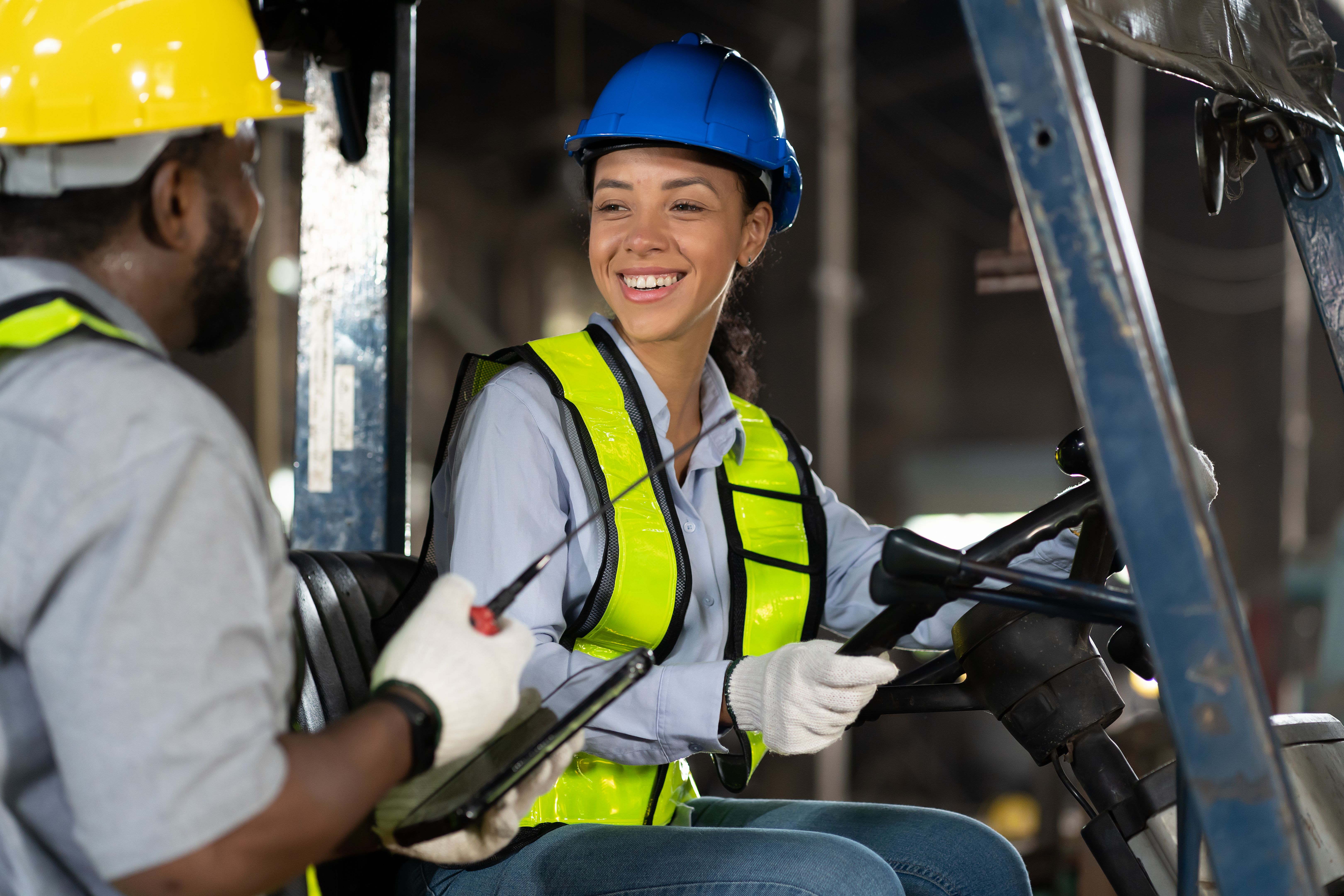 Happy female engineer driving and operating on forklift truck in the industry factory. Woman technician wear safety helmet, glasses and uniform working and driving forklift truck
