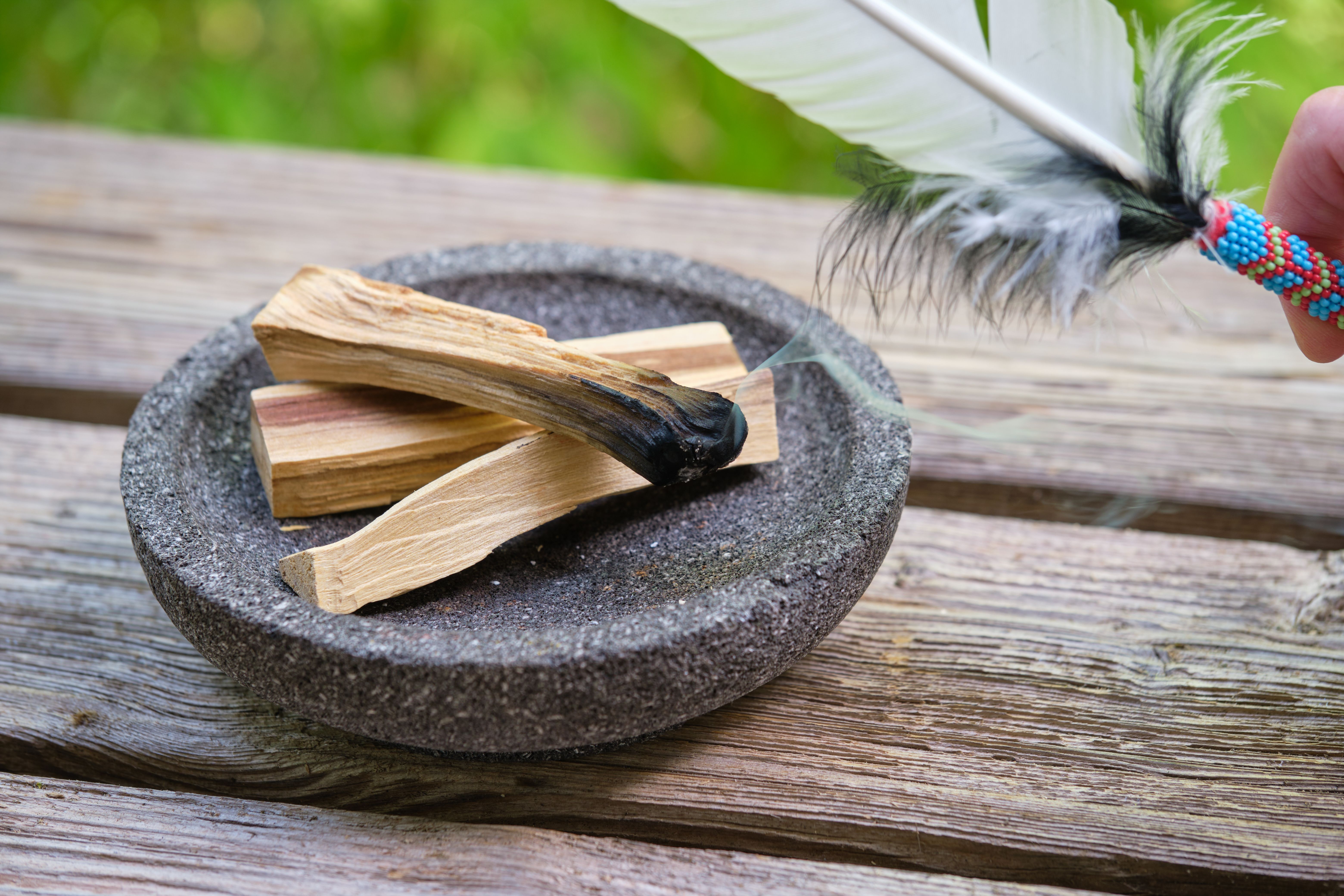 Burning palo santo sticks with smoke and smudging feather outdoors in the garden in summer on a wooden table