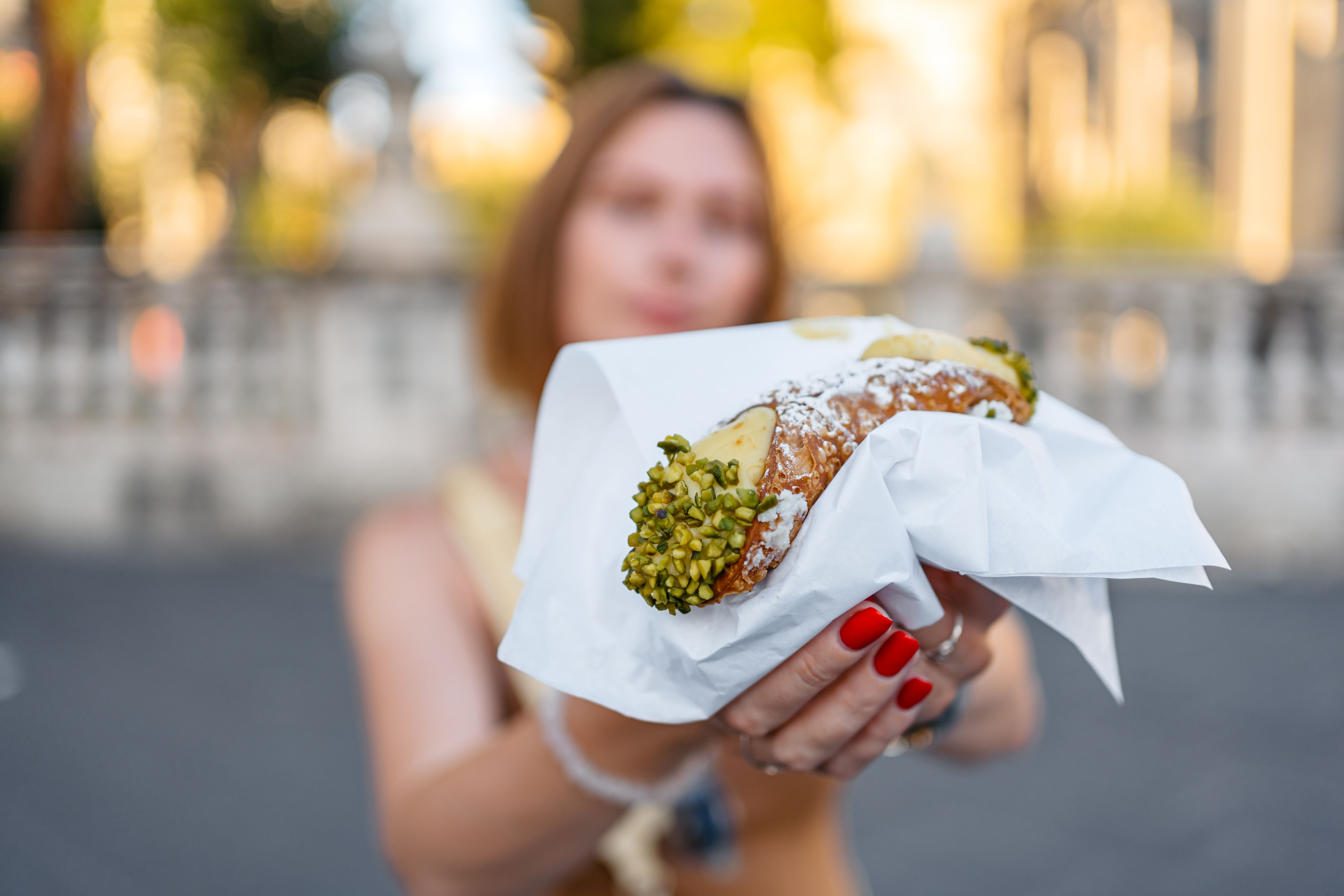 Mid-Adult Woman Eating A Cannoli Pastry Filled With Creamy Ricotta And Pistachios On The Street In Catania, Sicily Mid-Adult Woman Eating A Cannoli Pastry Filled With Creamy Ricotta And Pistachios On The Street In Catania, Sicily