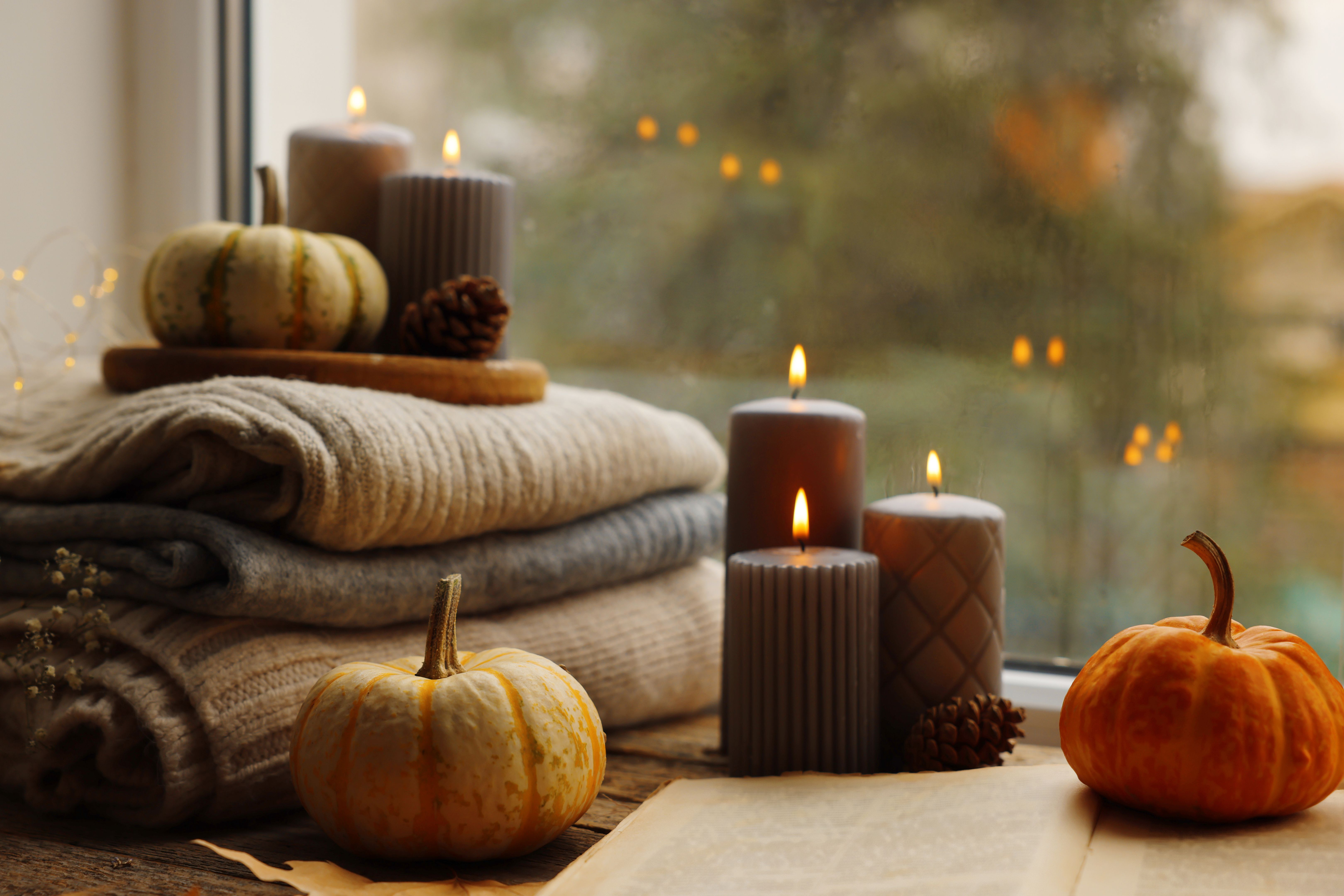 Burning candles, open book and stack of soft sweaters on windowsill. Autumn atmosphere