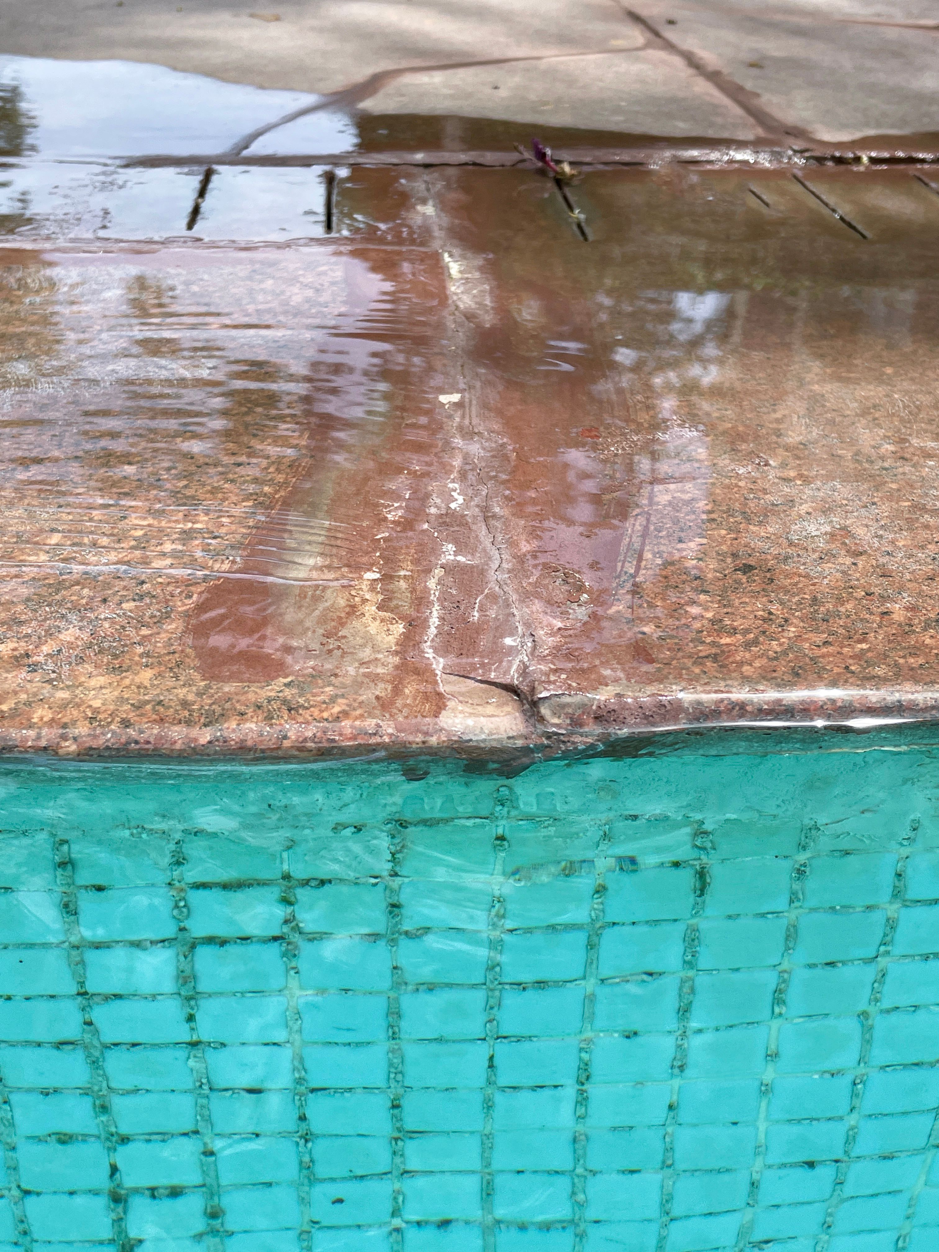 Close-up image of cracked, square slab of poolside stone patio, outdoor, swimming pool with turquoise blue mosaic tiles distorted by rippled water, puddle leak, splashed water, focus on foreground