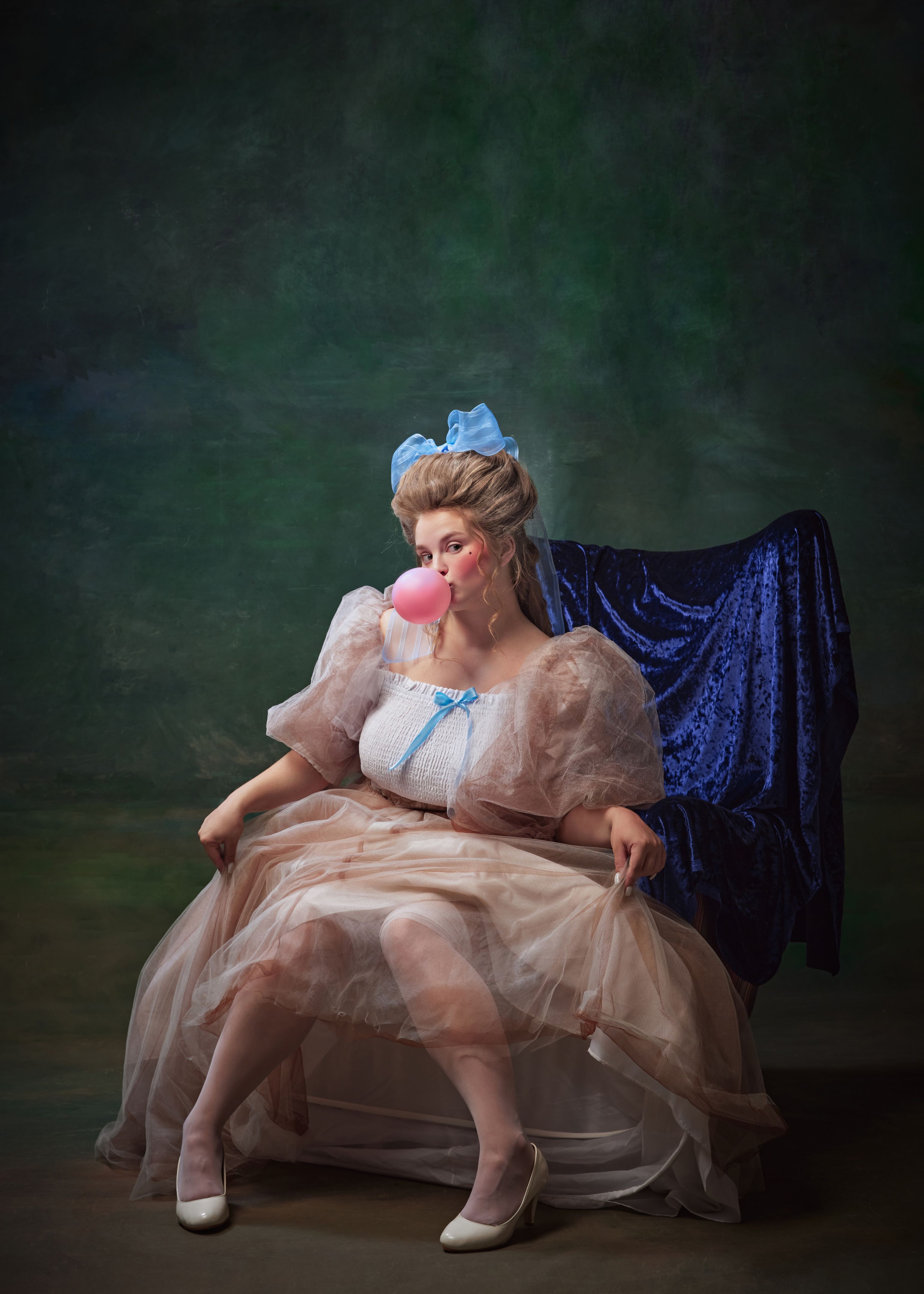 Beautiful woman in period attire, royal person sitting on chair with pink bubble gum against vintage green background