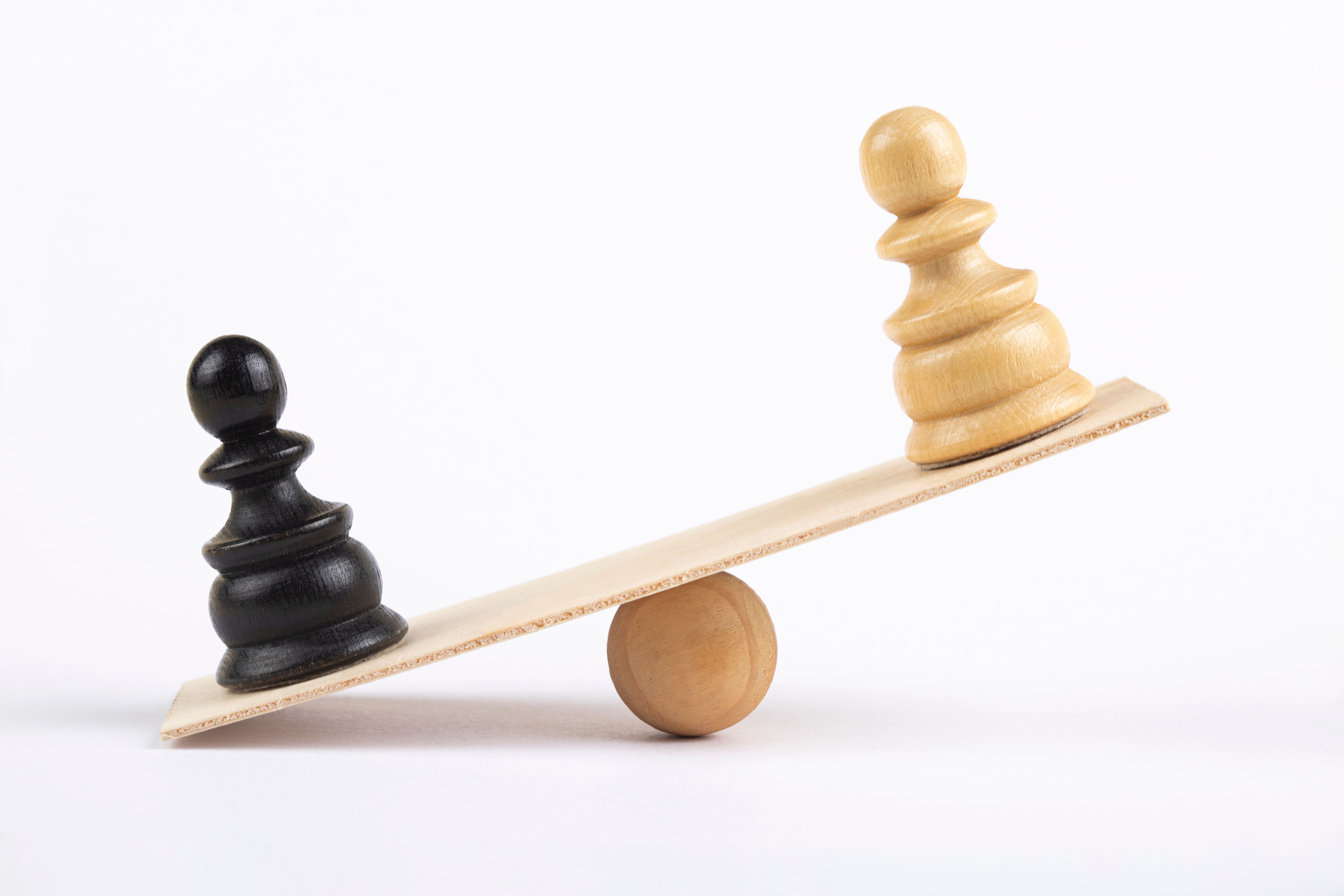 White and a black pawn chess piece stand on wooden seesaw
