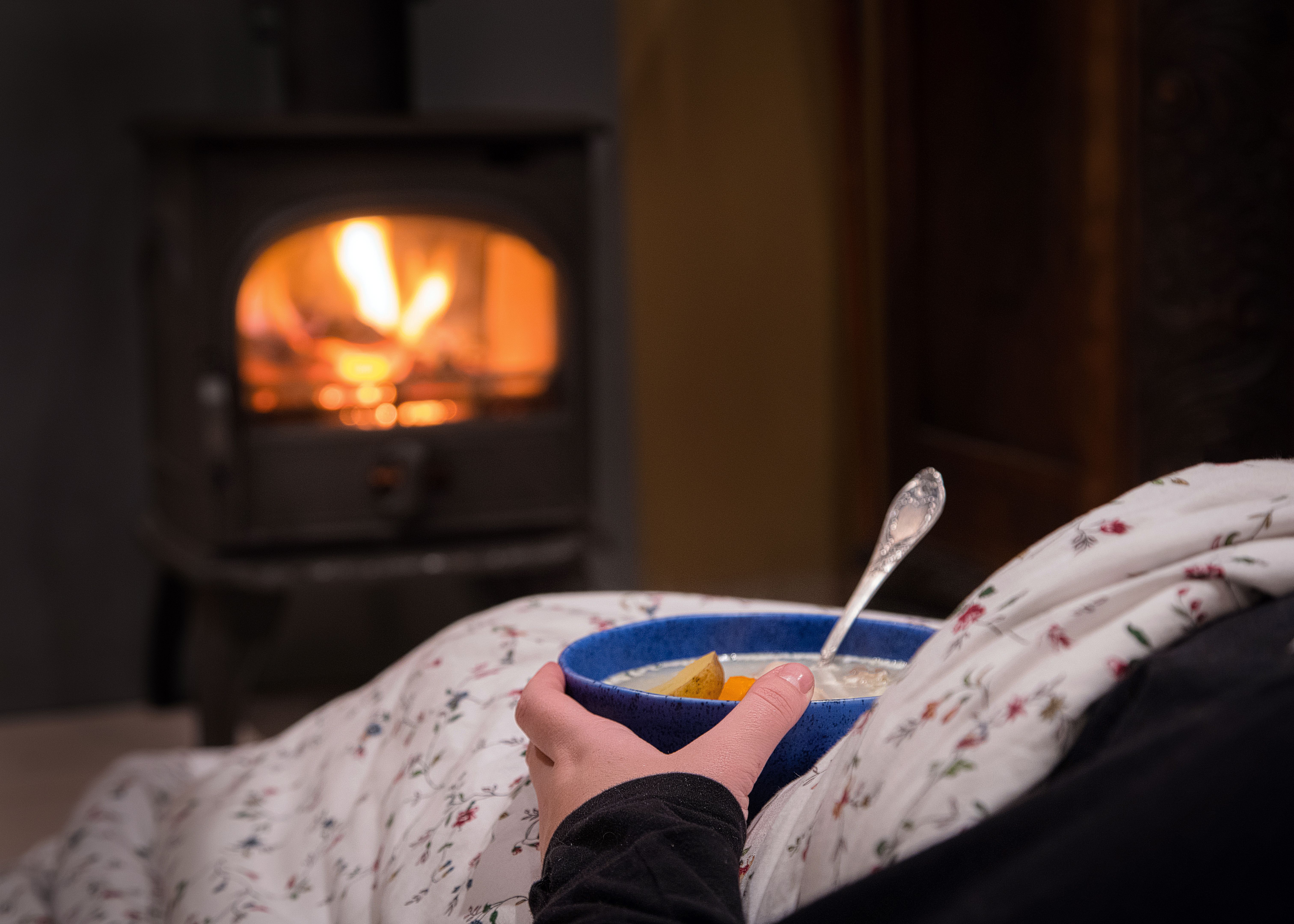 Woman having a hot bowl of vegetable soup at the fireplace. Burning fireplace with wooden logs burning inside in the background. Warm light, romantic, Christmas like atmosphere