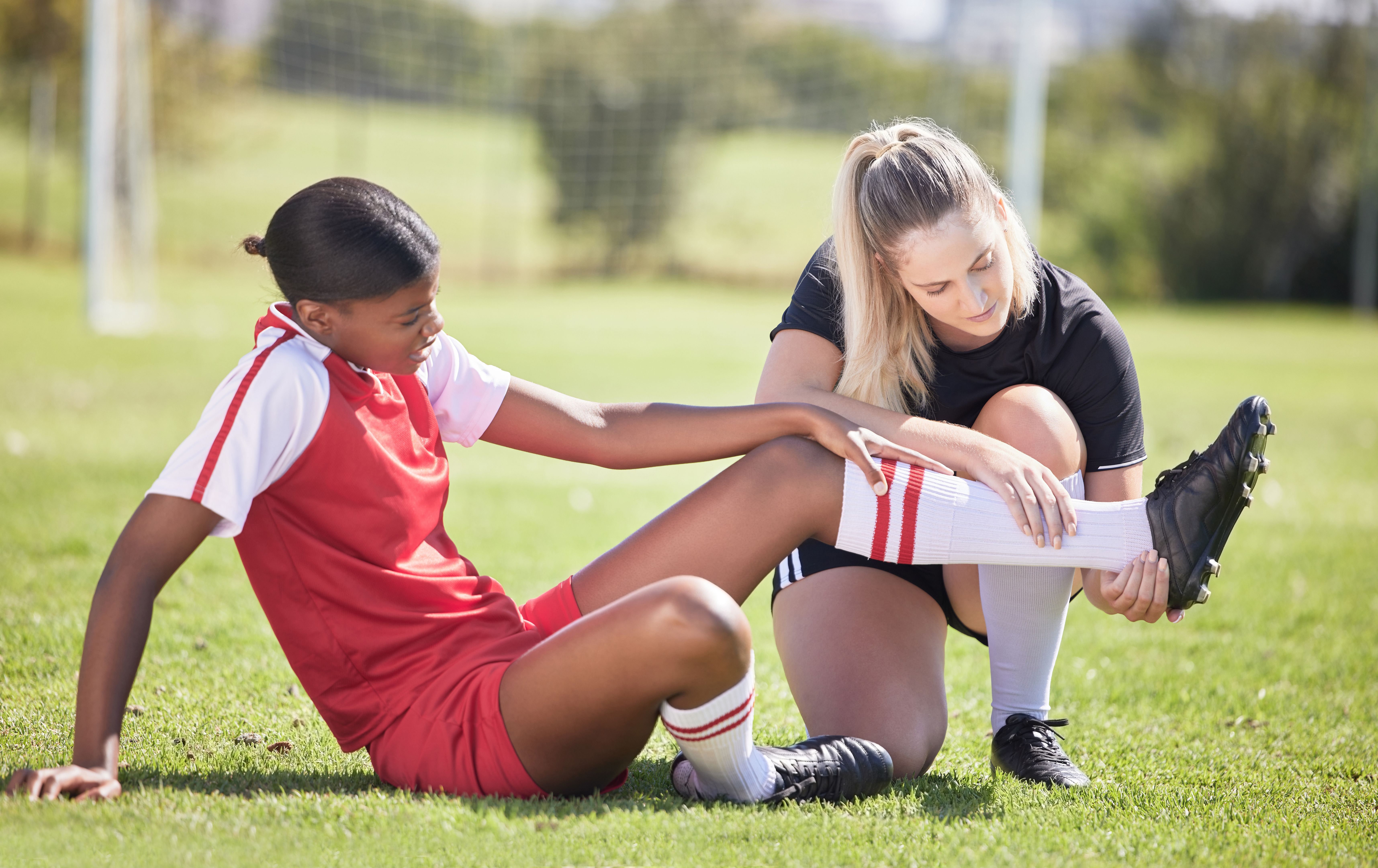 Soccer, sports and injury of a female player suffering with sore leg, foot or ankle on the field. Painful, hurt and discomfort woman getting her pain checked out by athletic trainer on the pitch.