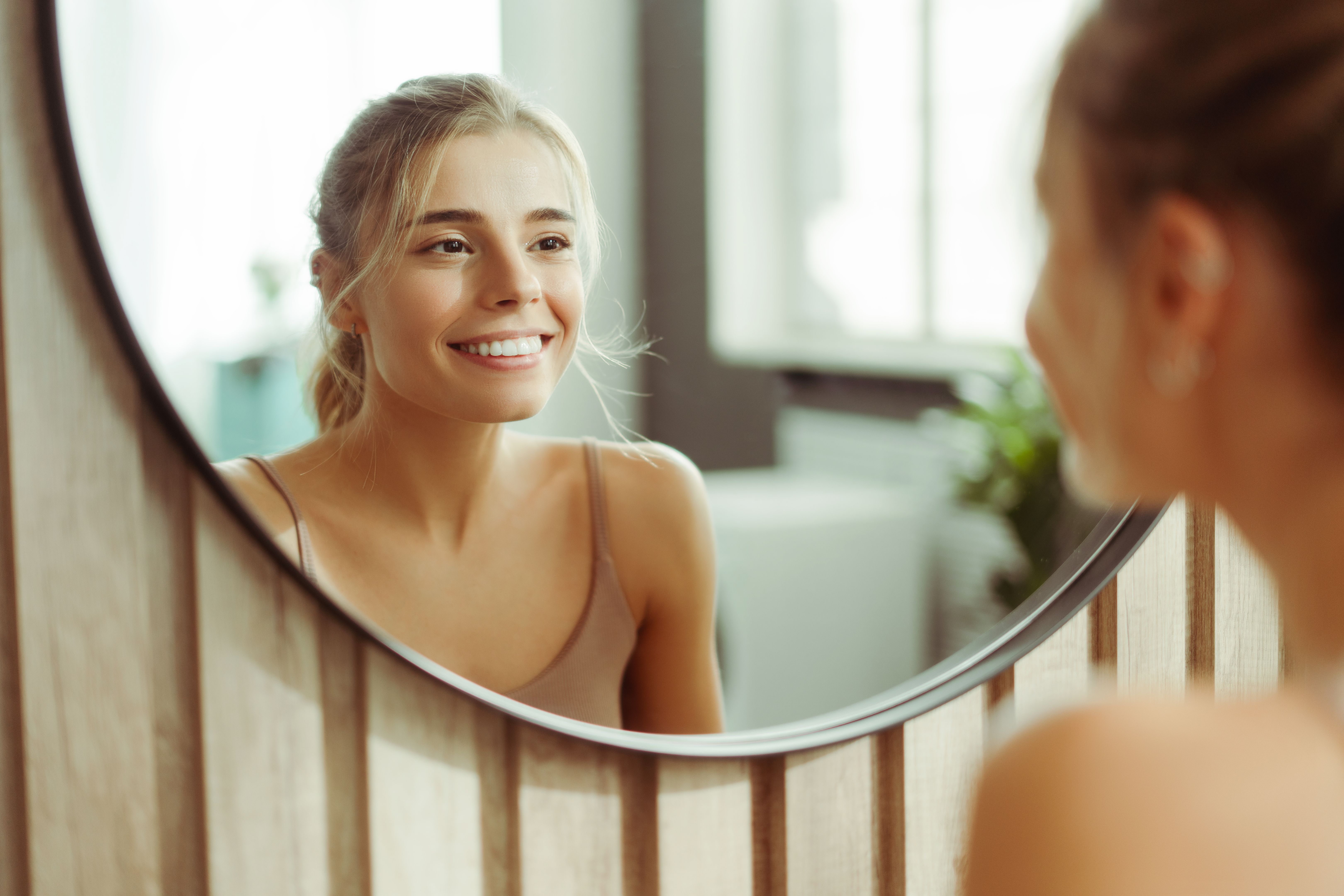 Smiling, confident woman with make up wearing tank top, looking in mirror, standing in bathroom