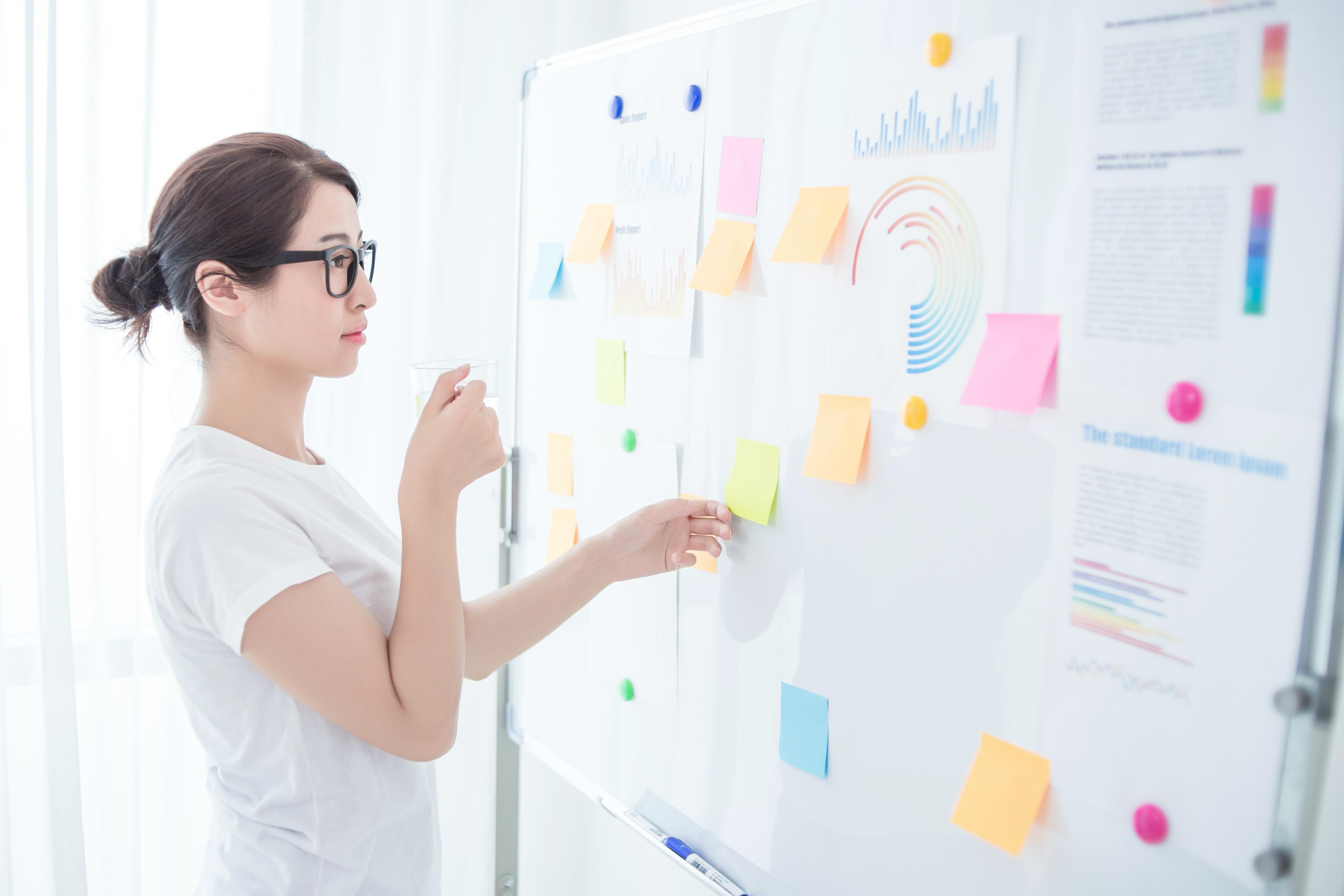Asian woman in glasses standing by a whiteboard with sticky notes and holding a cup Asian woman in glasses standing by a whiteboard with sticky notes and holding a cup