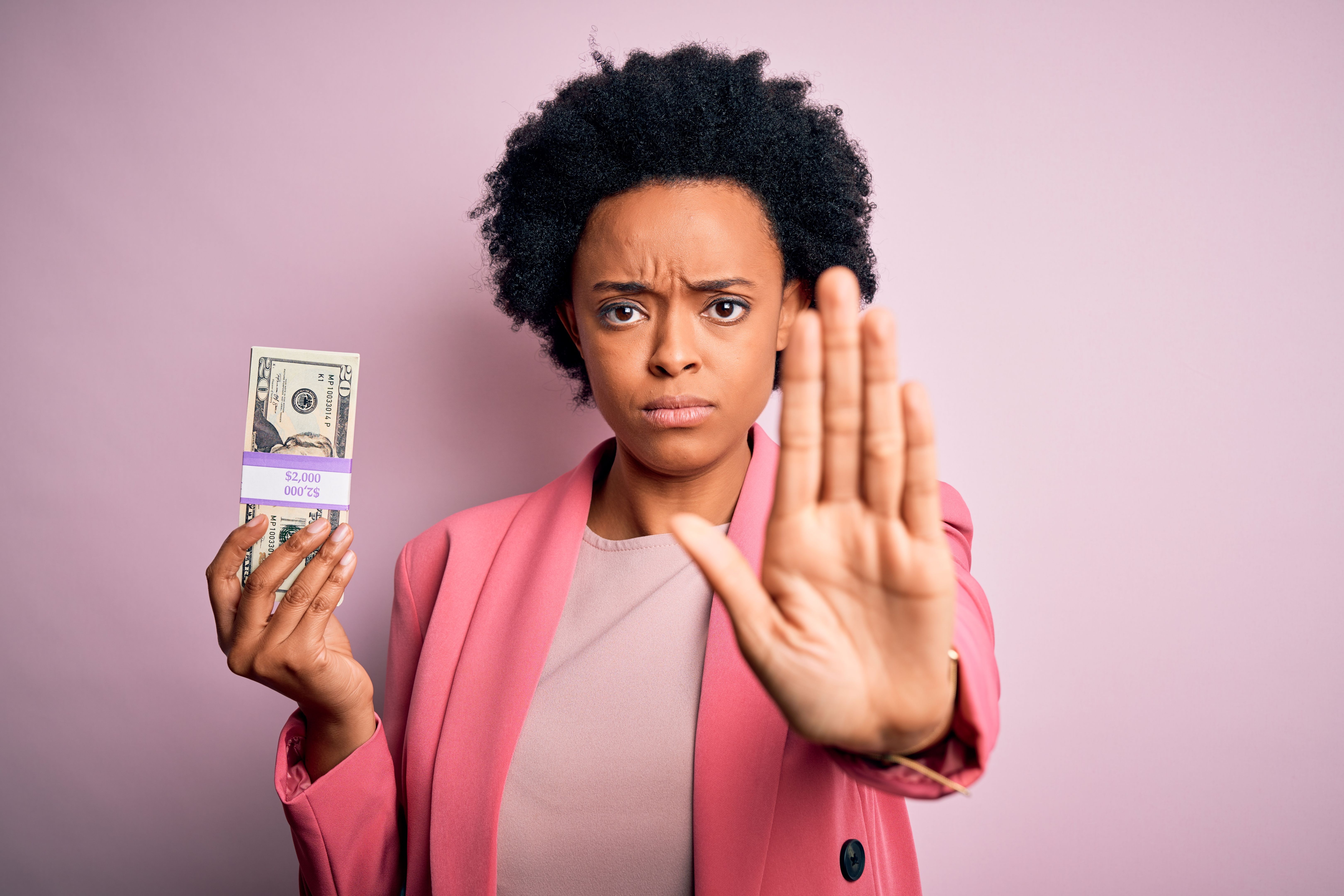 Young African American afro businesswoman with curly hair holding dollars banknotes with open hand doing stop sign with serious and confident expression, defense gesture