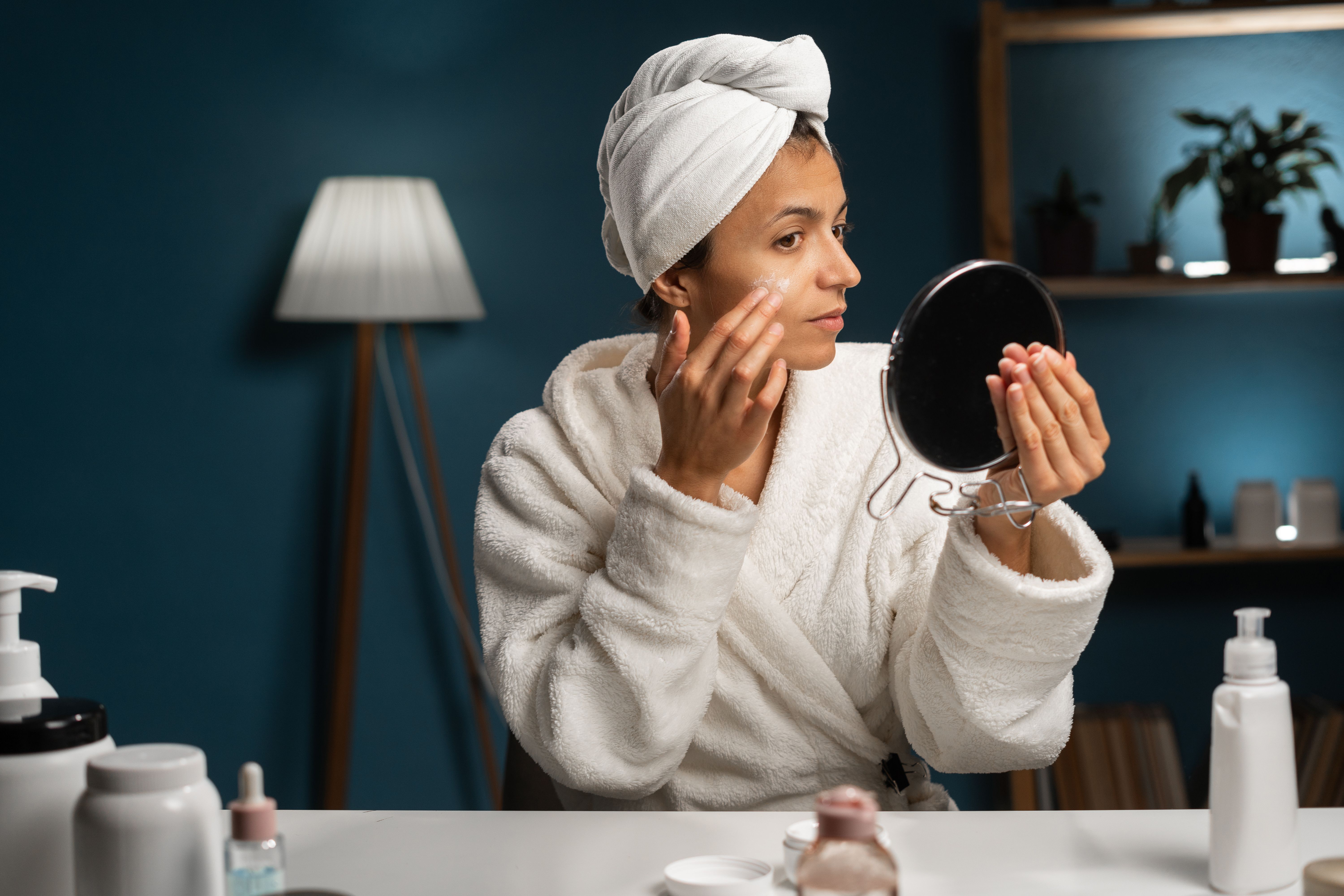 Beautiful hispanic woman applying face cream sitting at the table. Smiling young woman in bathrobe doing skin care routine at home.