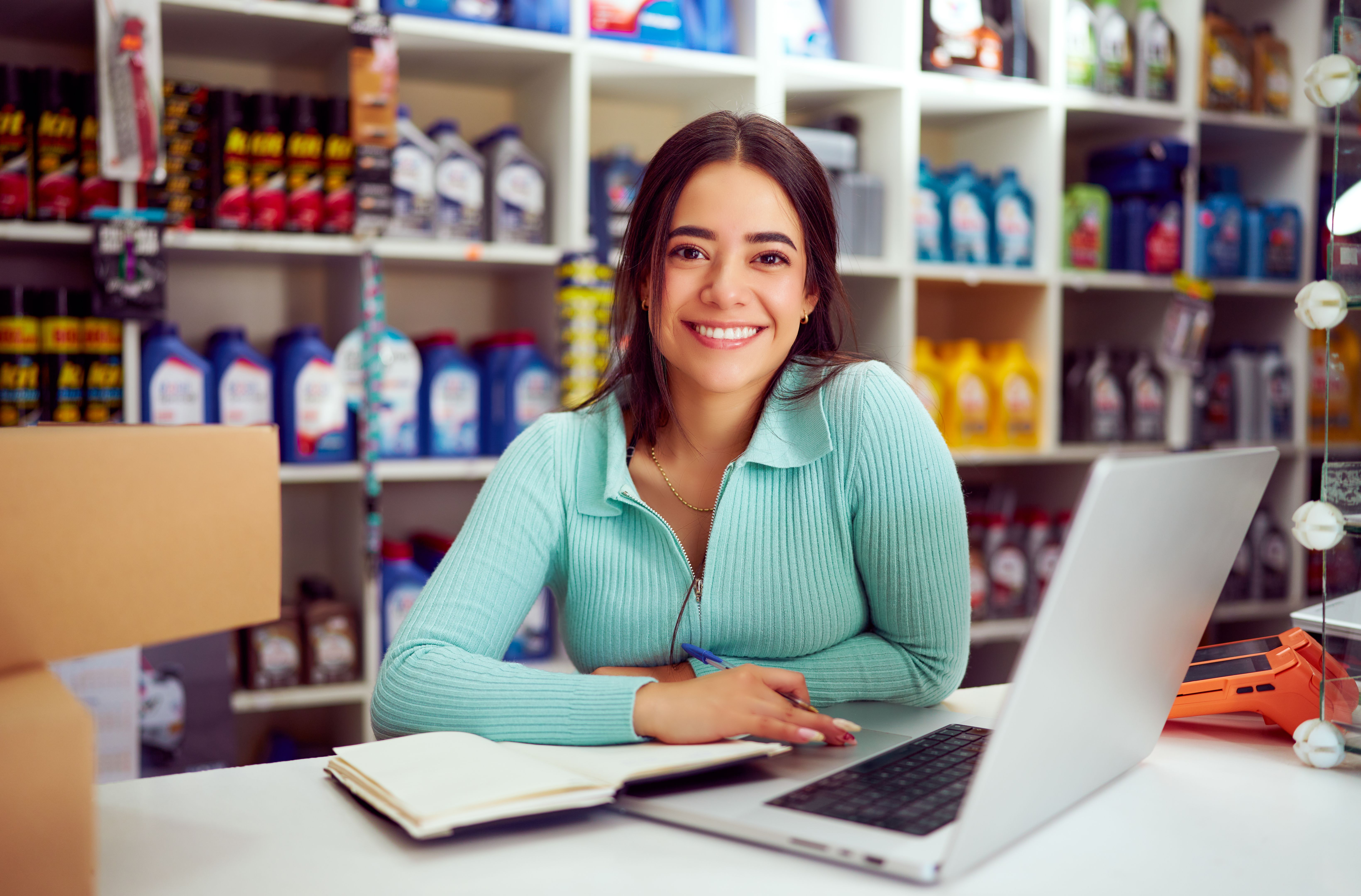 Jeune vendeuse latine souriante et travaillant sur un ordinateur portable dans un magasin de pièces automobiles Jeune vendeuse latine souriante et travaillant sur un ordinateur portable dans un magasin de pièces automobiles