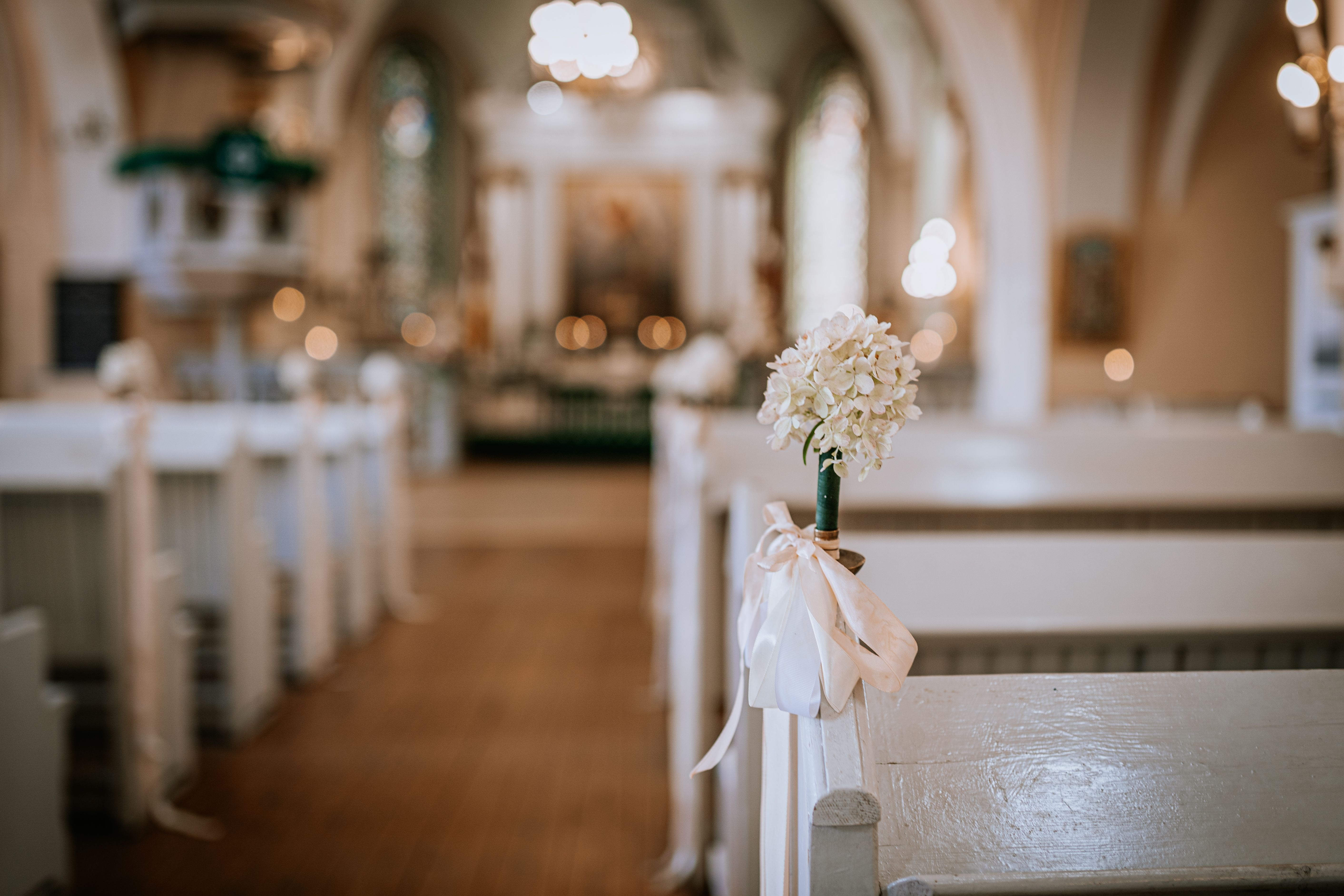 Close-up of a white floral wedding decoration with ribbon on a church pew, with a blurred altar and soft lighting in the background.