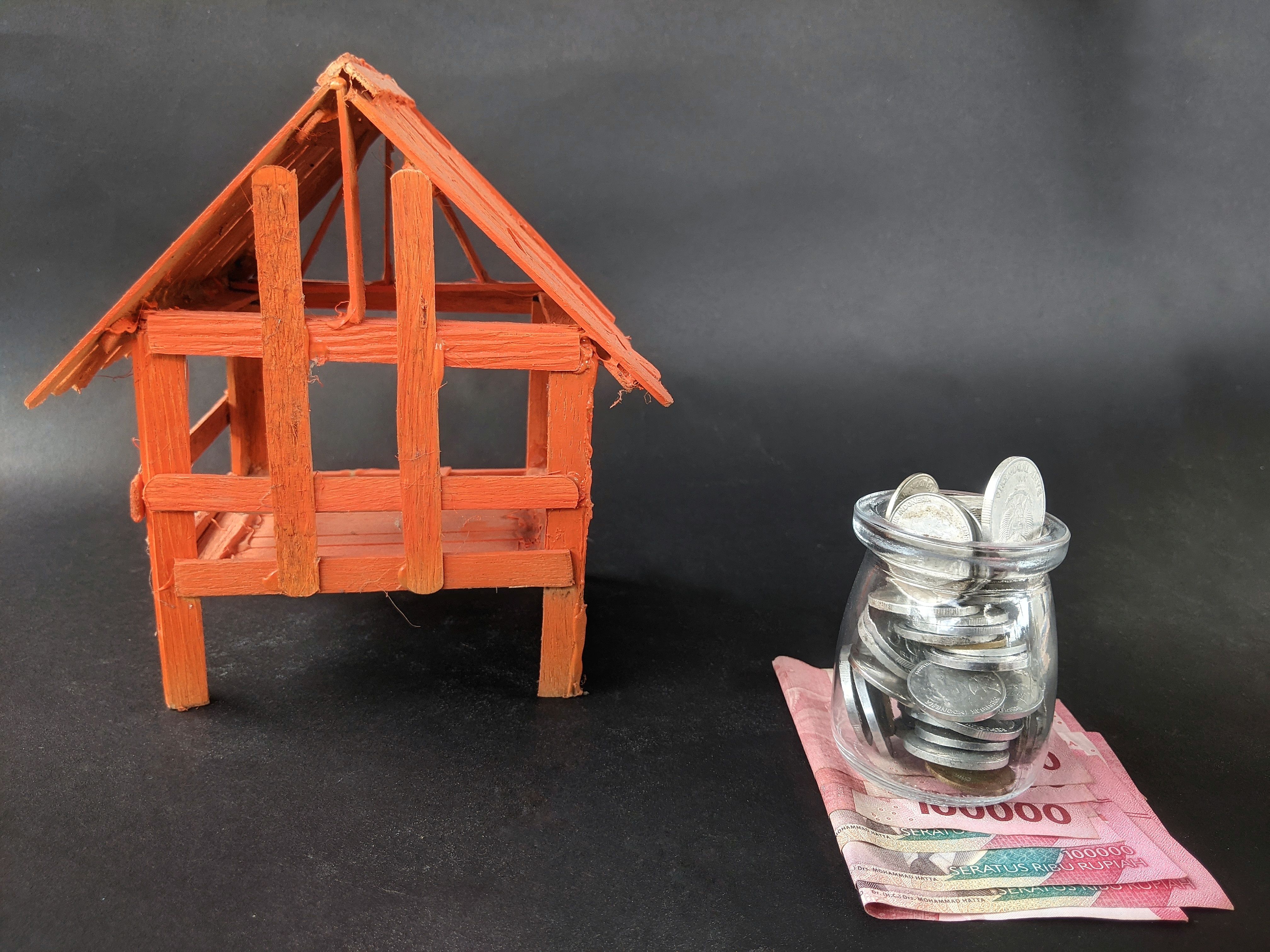 A miniature orange wooden house or shelter hut model is placed next to Indonesian Rupiah banknotes and coins on a black background.