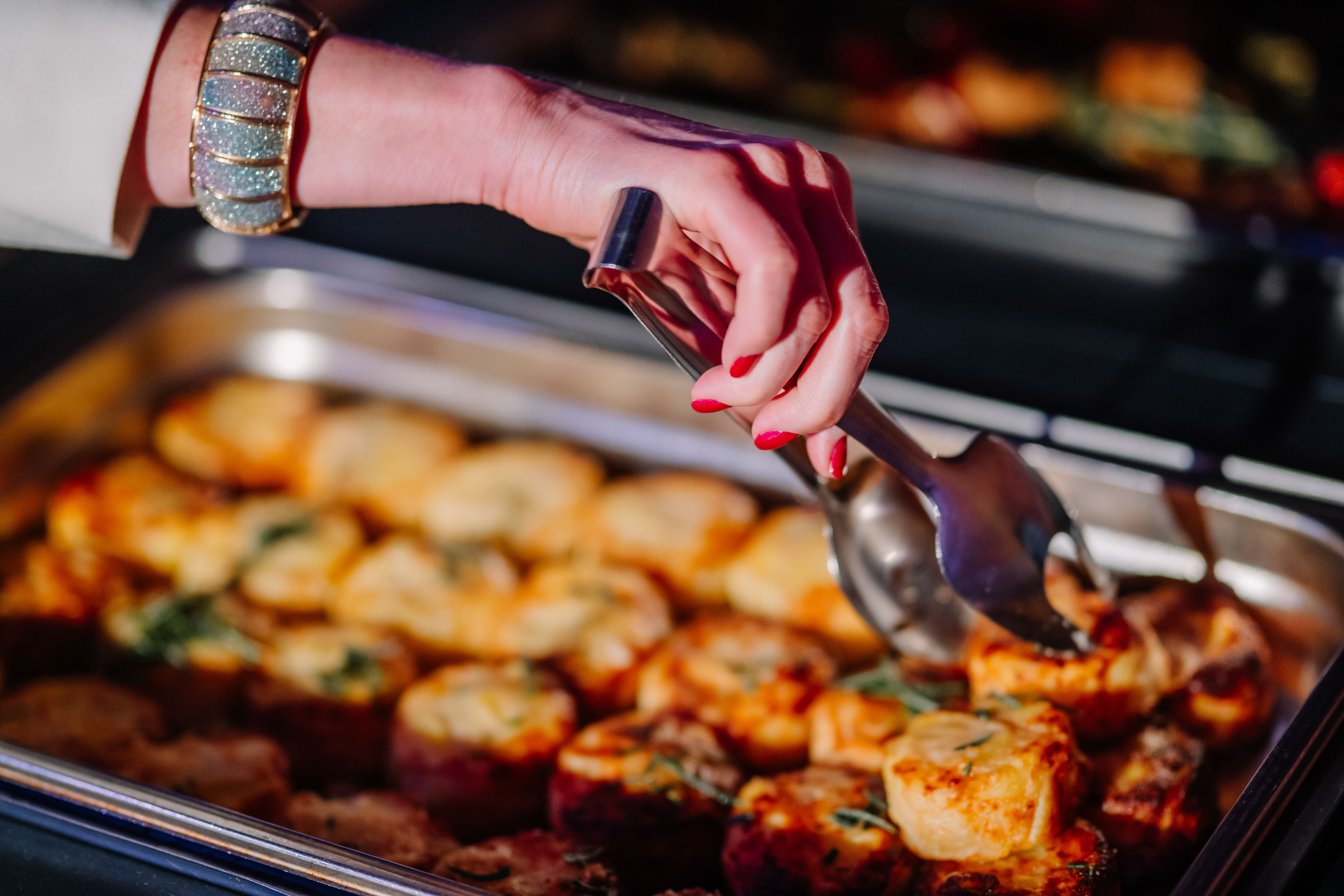 Close-up of a hand with red nails and bracelet using tongs to serve golden roasted potatoes from a buffet tray during a catered event. Close-up of a hand with red nails and bracelet using tongs to serve golden roasted potatoes from a buffet tray during a catered event.