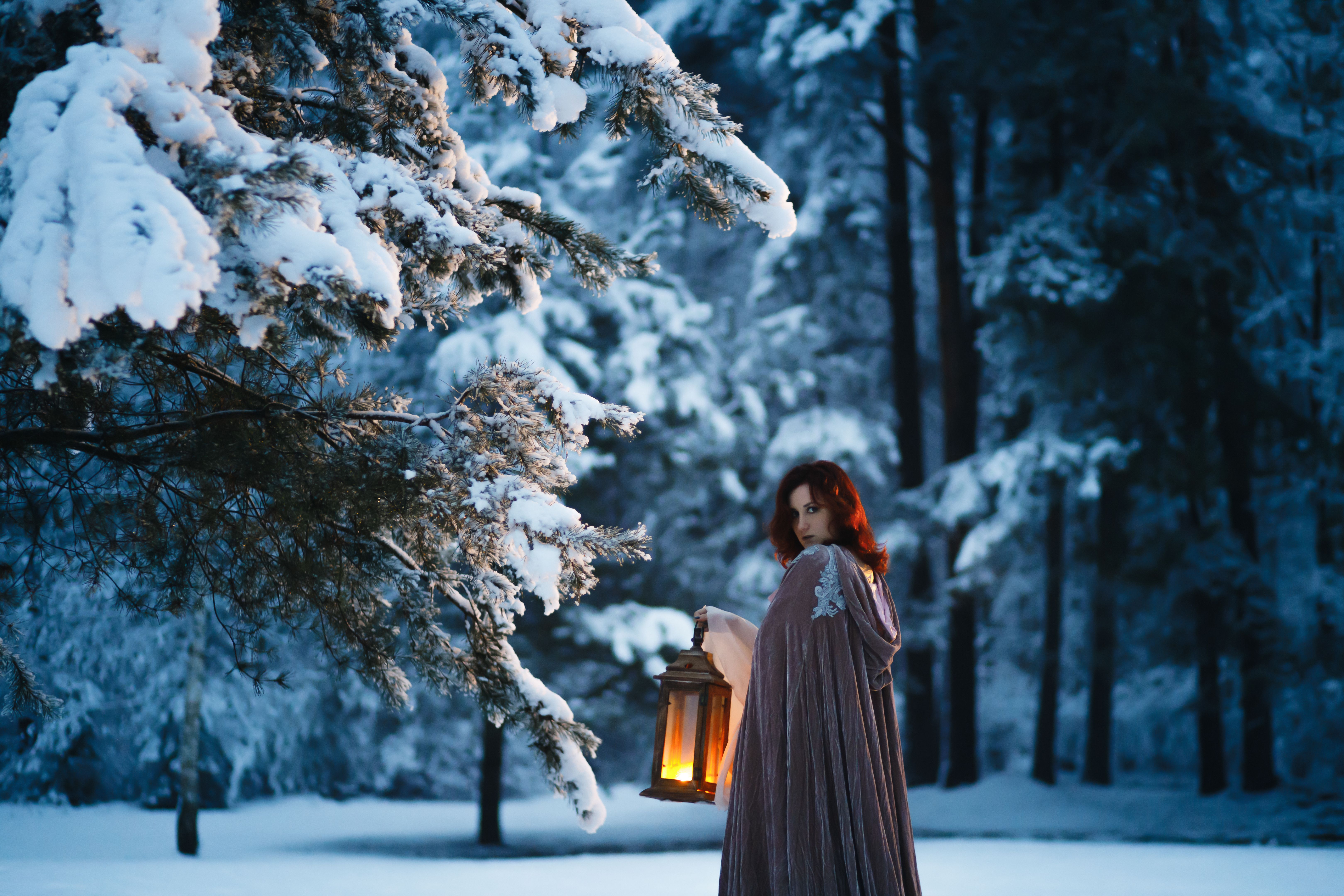Fantasy cosplay: Young woman with vintage lantern looking at fir tree covered in snow. Medieval fairytale concept