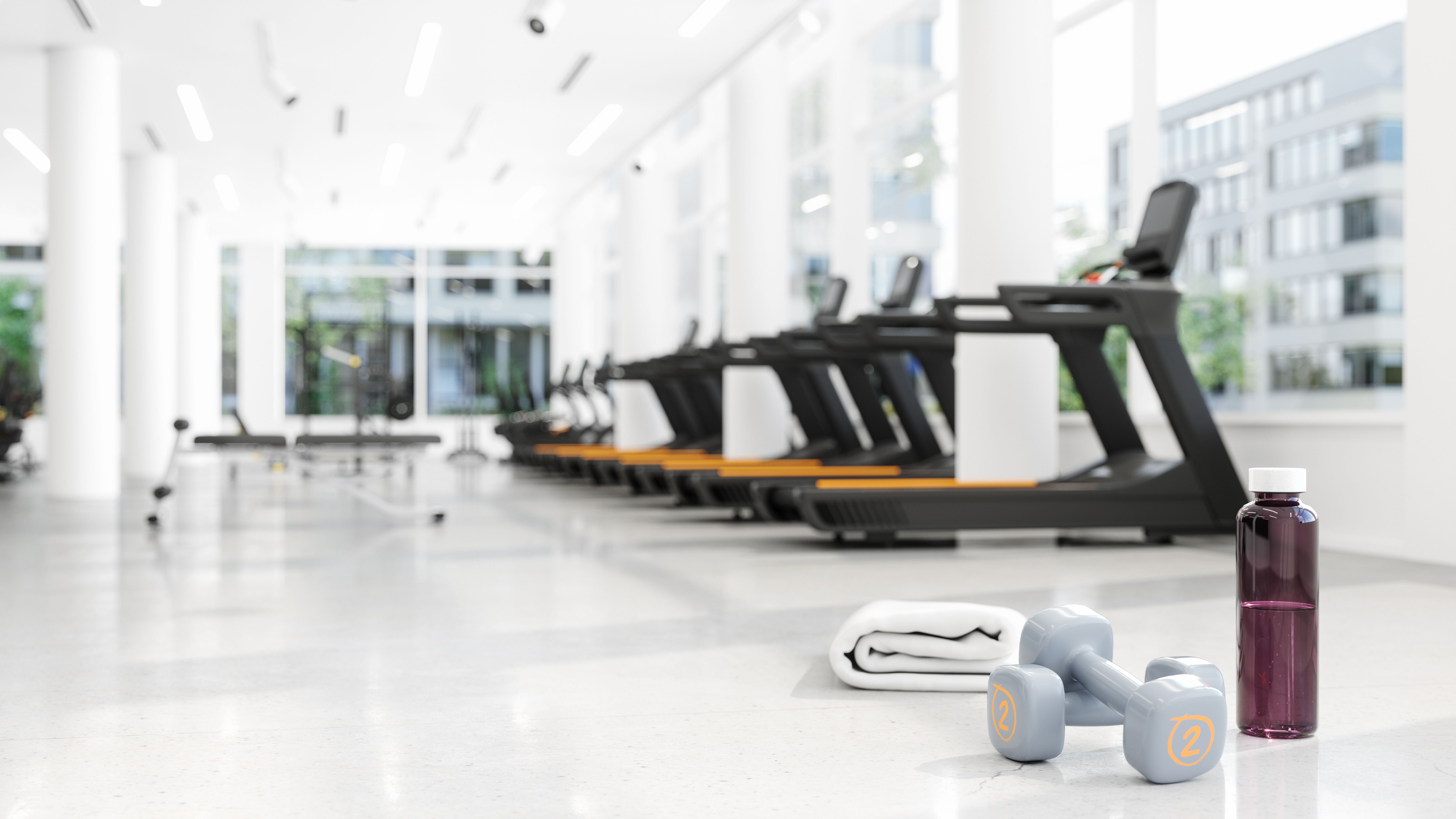 Close-up View Of Water Bottle, Towel And Dumbbells On The Floor With Blurred Gym Background