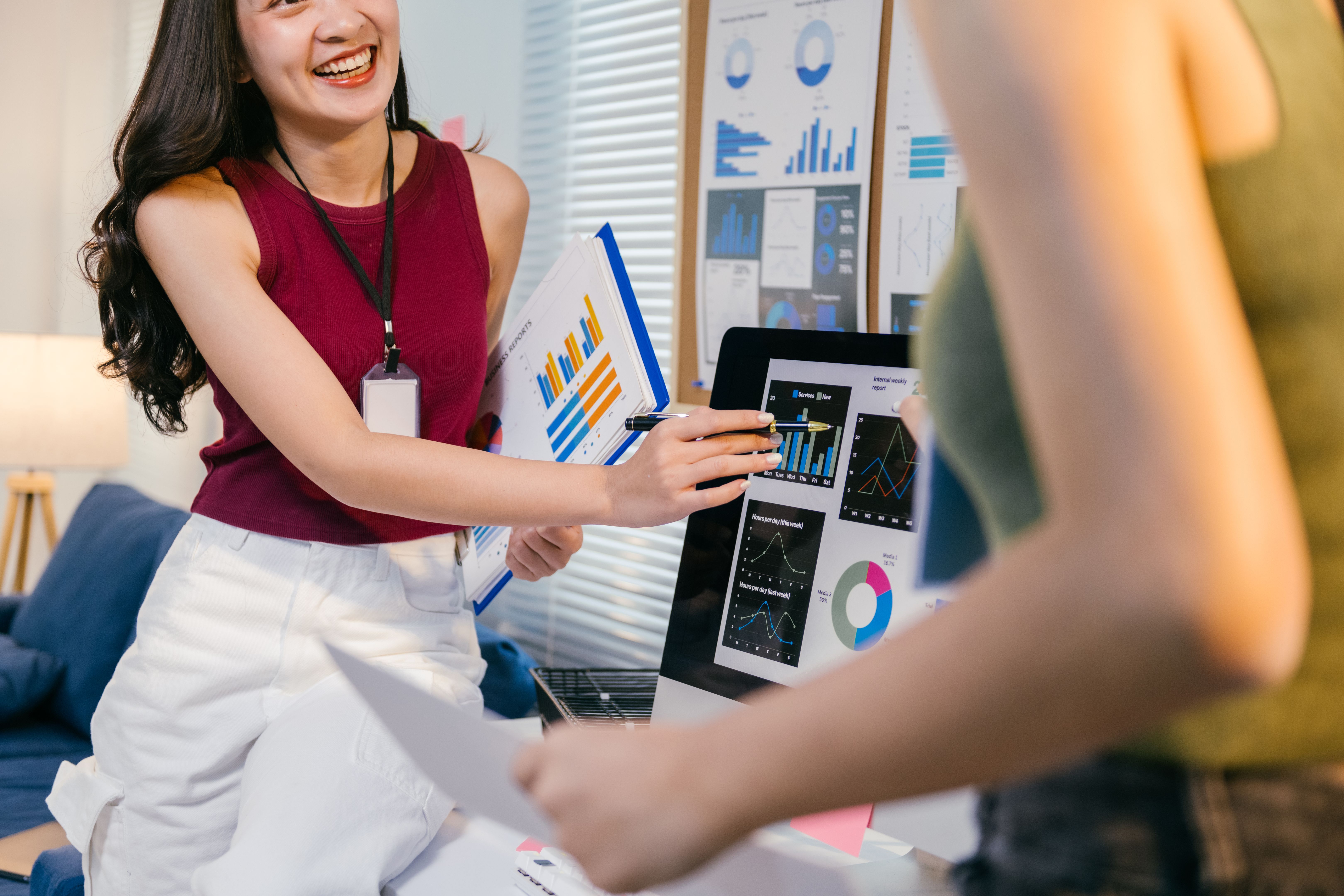 Two young businesswomen analyzing charts on a computer screen, discussing financial results while collaborating in a modern office environment with printed reports and a pen