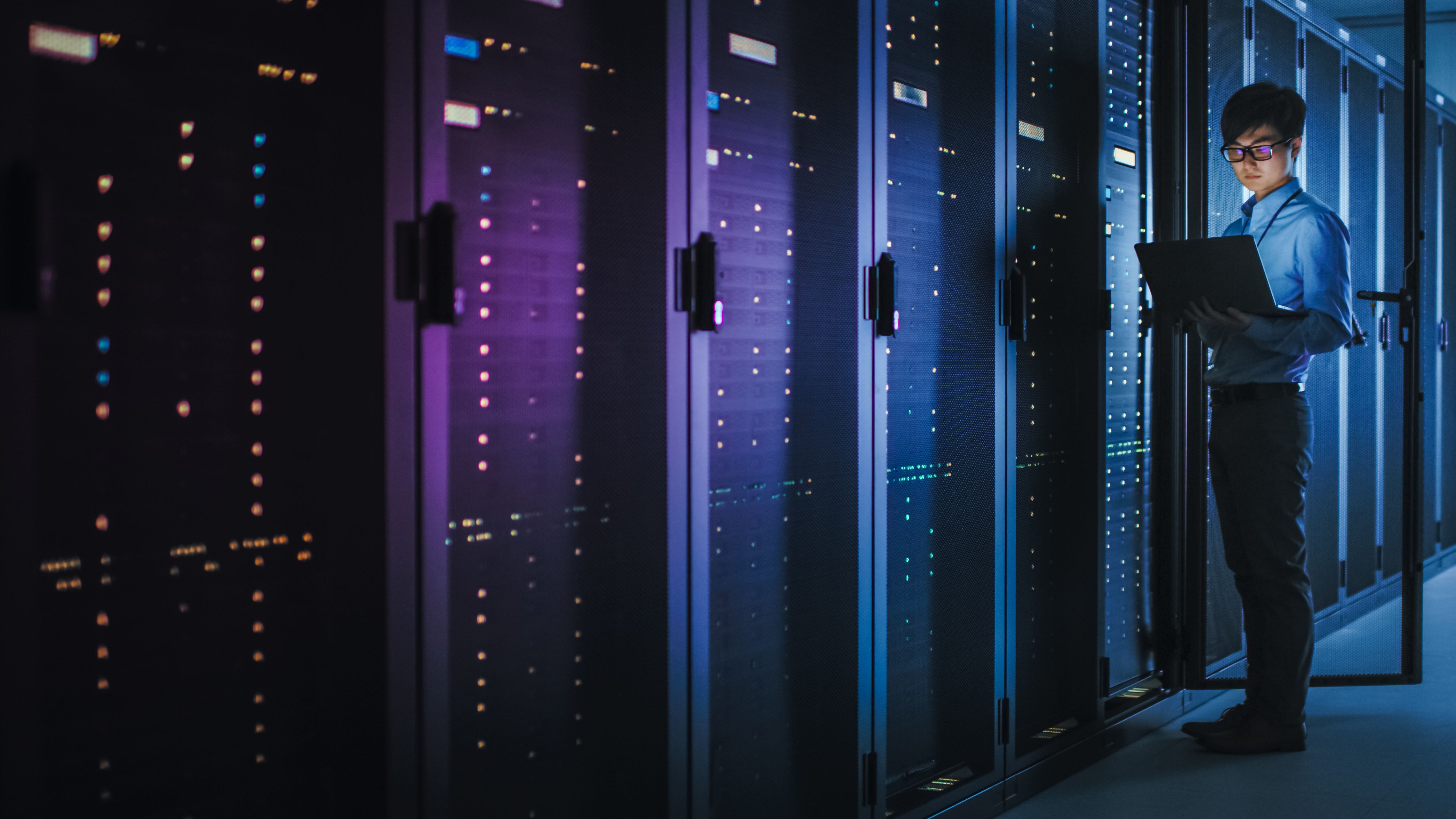 In Dark Data Center:  IT Specialist Stands Beside the Row of Operational Server Racks, Uses Laptop for Maintenance. Concept for Cloud Computing, Artificial Intelligence, Supercomputer, Cybersecurity. Neon Lights helps with the cooling for the environment