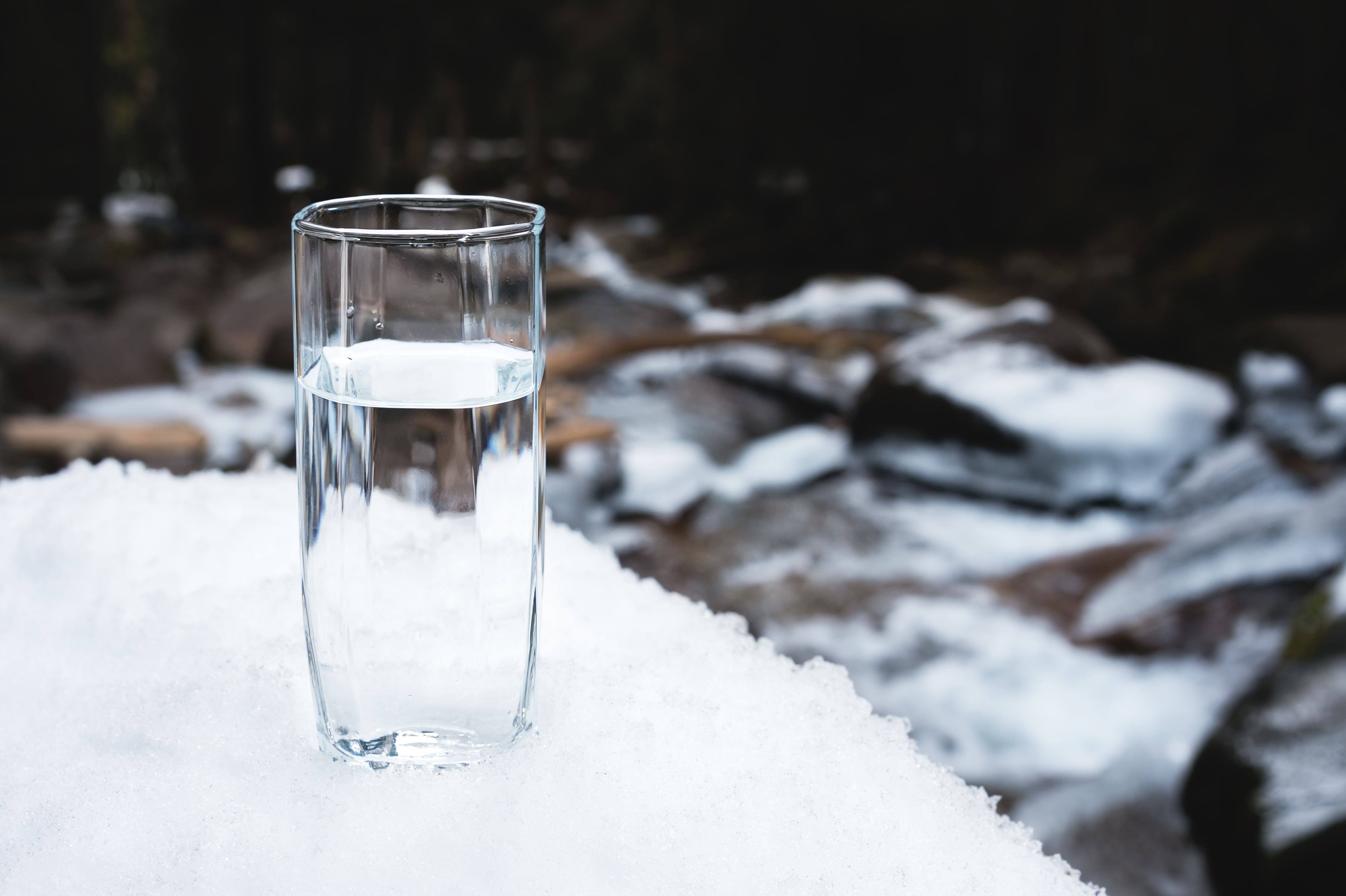 A transparent glass glass with drinking mountain water stands in the snow against a background of a clean mountain river and a forest in winter