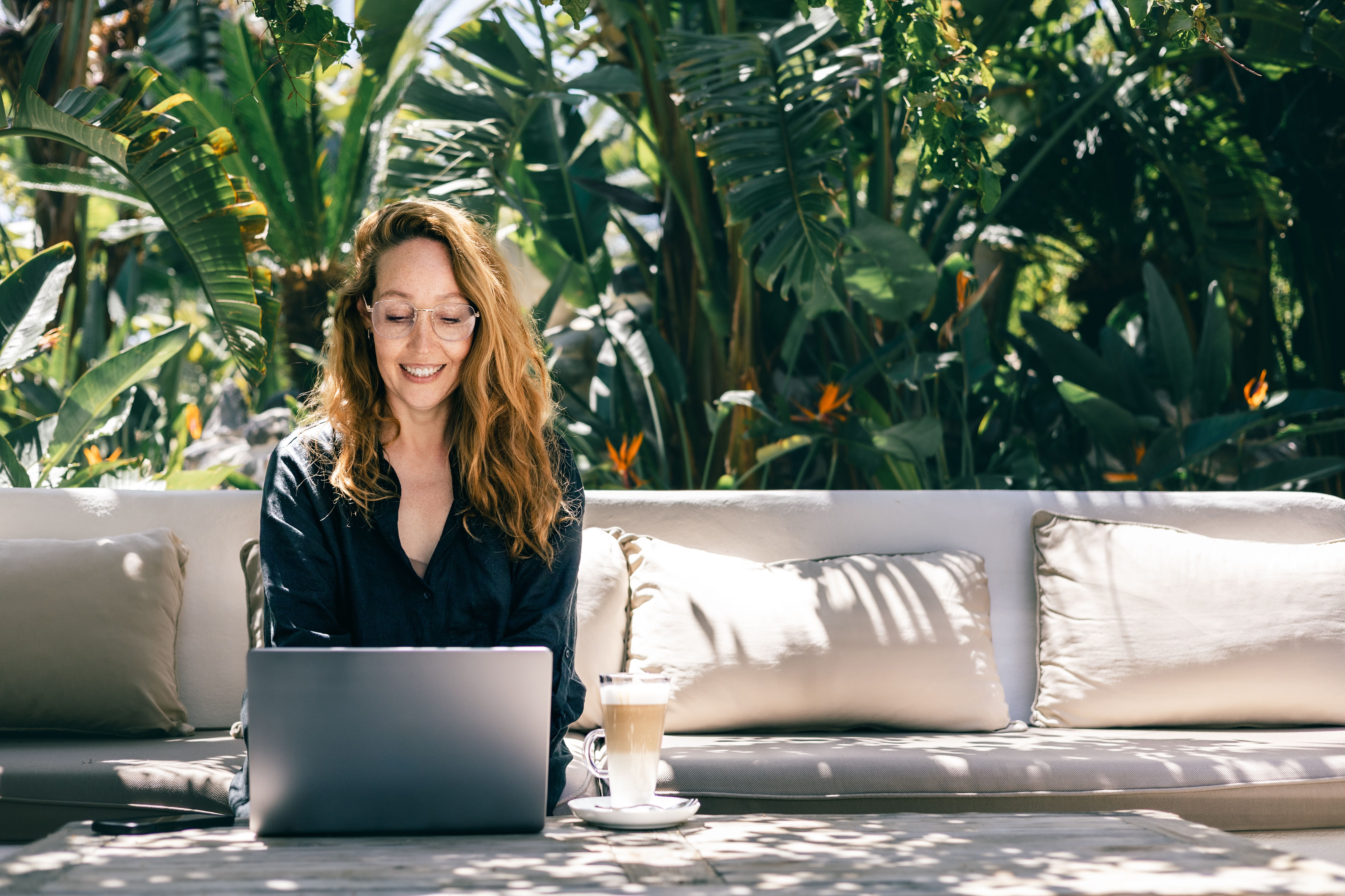 Smiling Woman Working on Laptop Outdoors in Tropical Garden with Coffee on Rustic Table