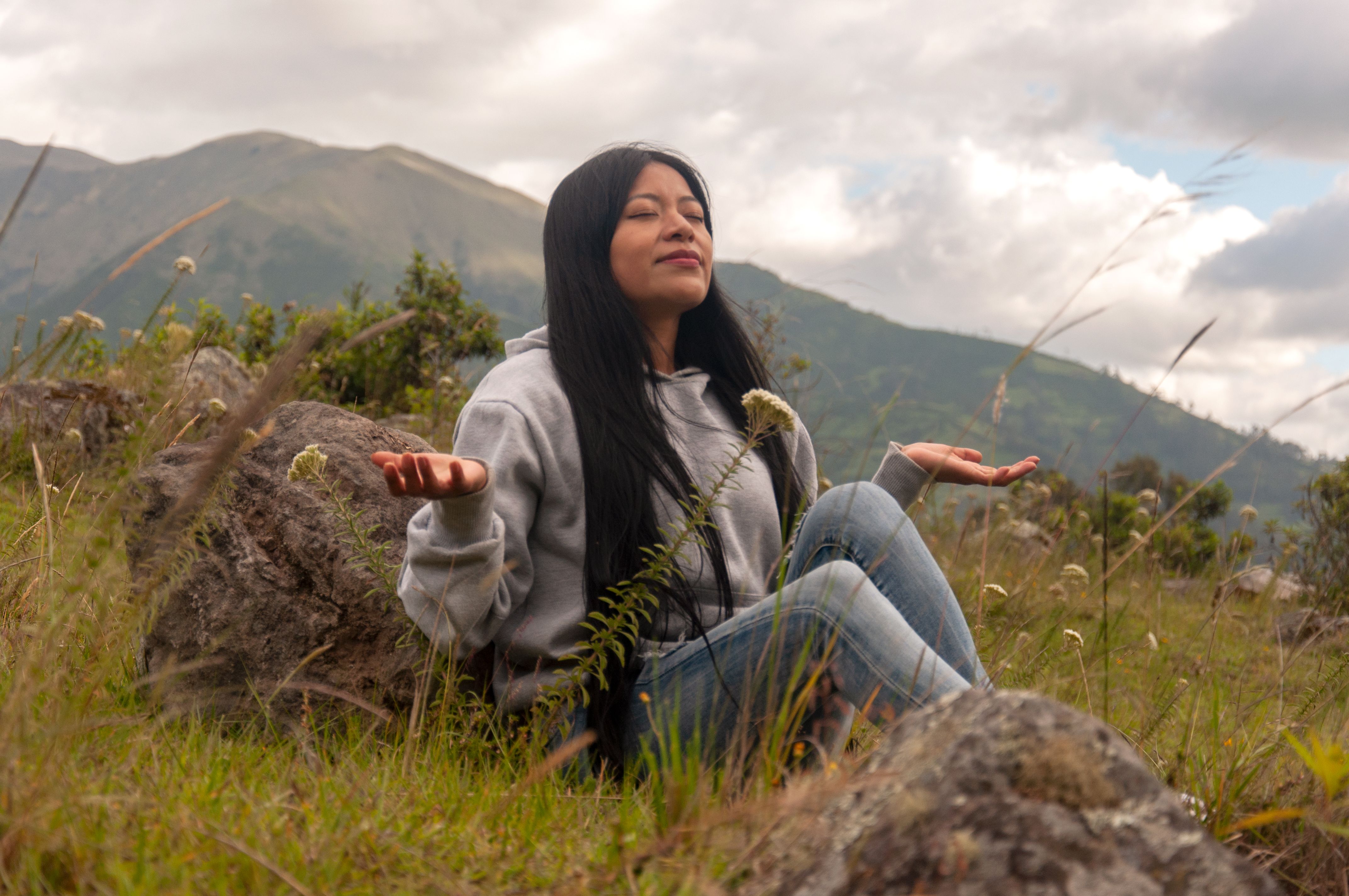 Una mujer está sentada con las piernas cruzadas en una ladera cubierta de hierba, meditando con los ojos cerrados, rodeada de flores primaverales