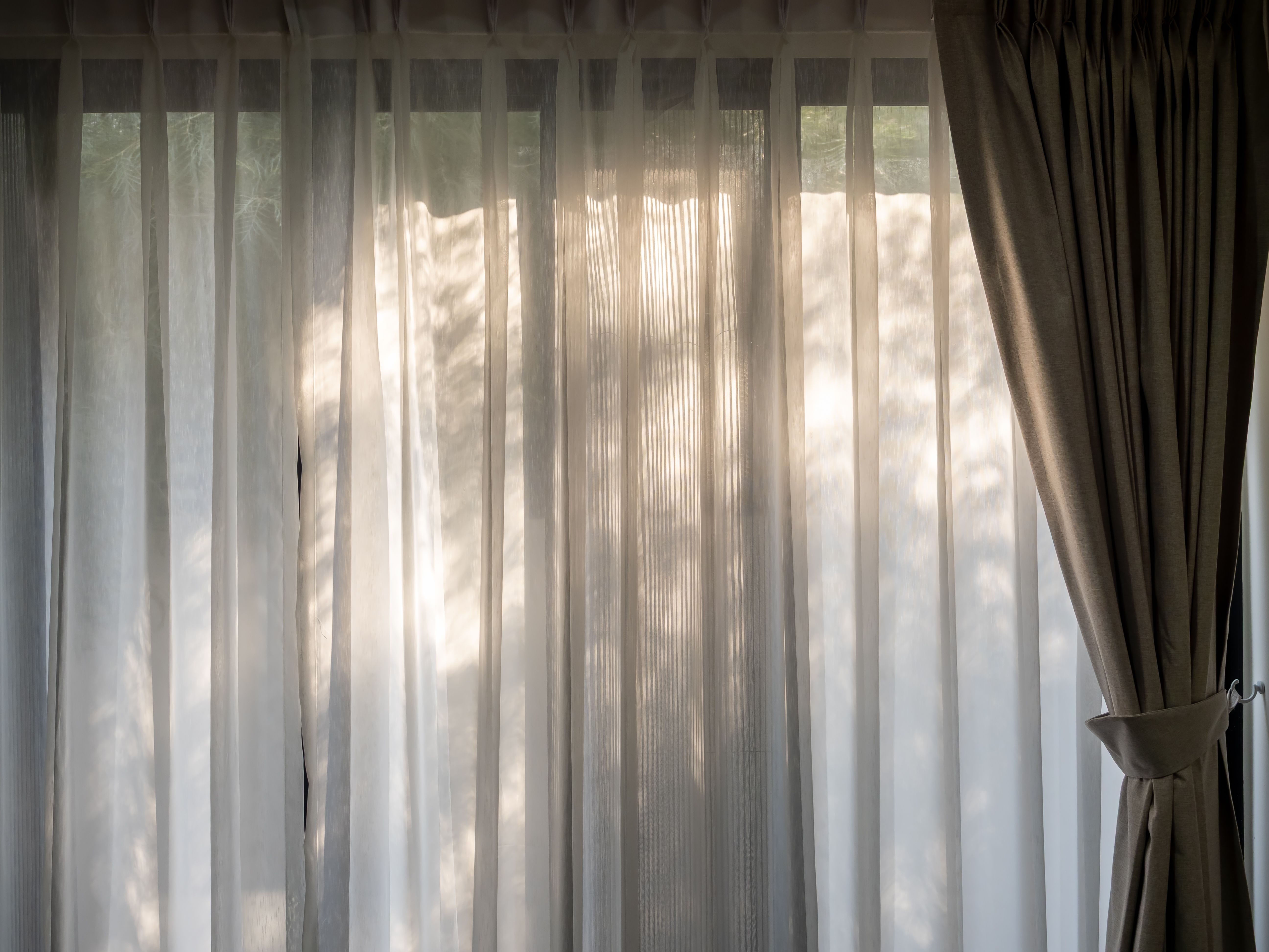 White transparent curtain at the window in the dark room with warm sunlight and tree leaves shade. Large horizontal window with closed white fabric curtain. Relaxation background concept. White transparent curtain at the window in the dark room with warm sunlight and tree leaves shade. Large horizontal window with closed white fabric curtain. Relaxation background concept.
