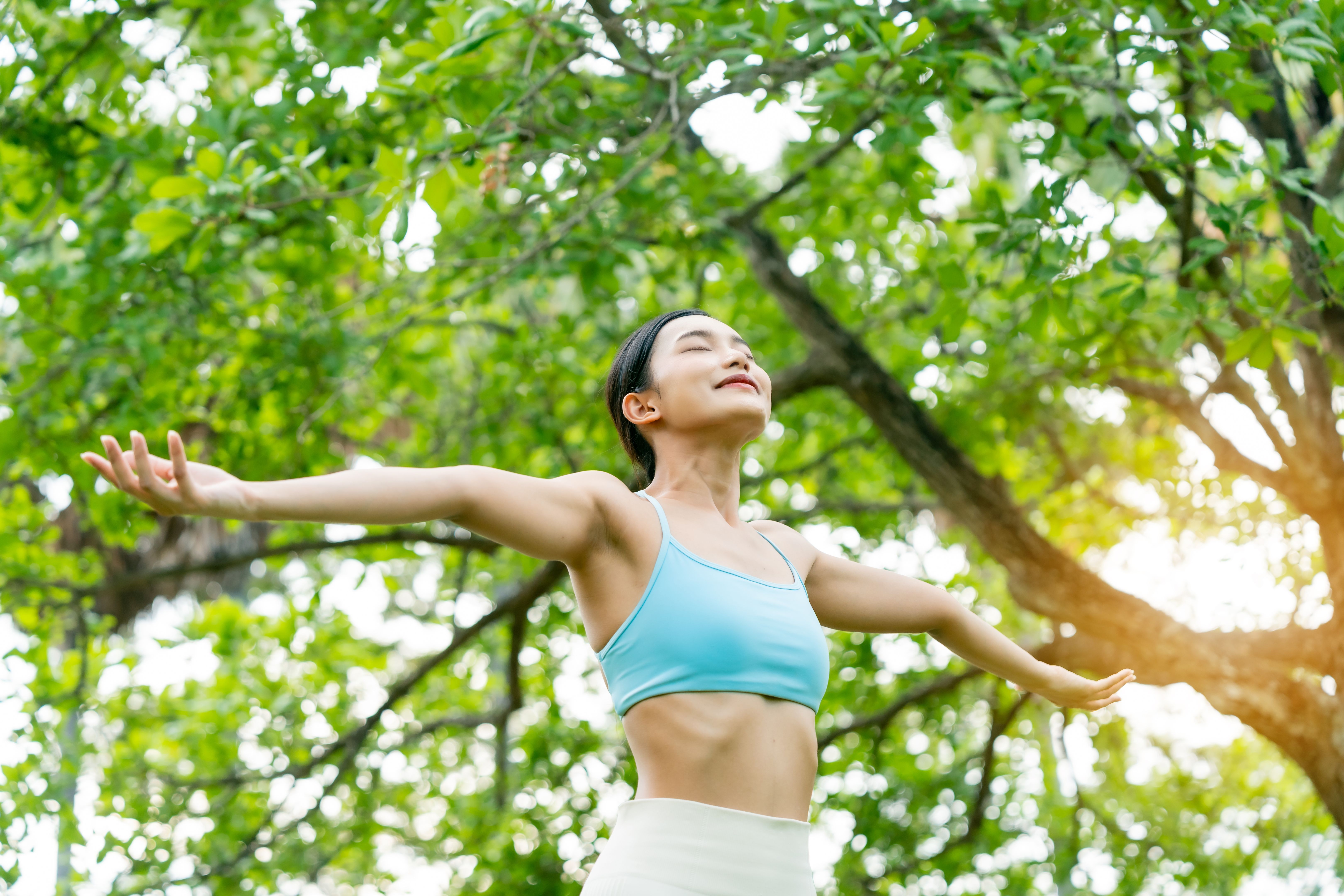 Young Asian woman relaxed breathing fresh air with trees in the background on a sunny day. Concept of wellness, healthy lifestyle, mindfulness, and outdoor fitness