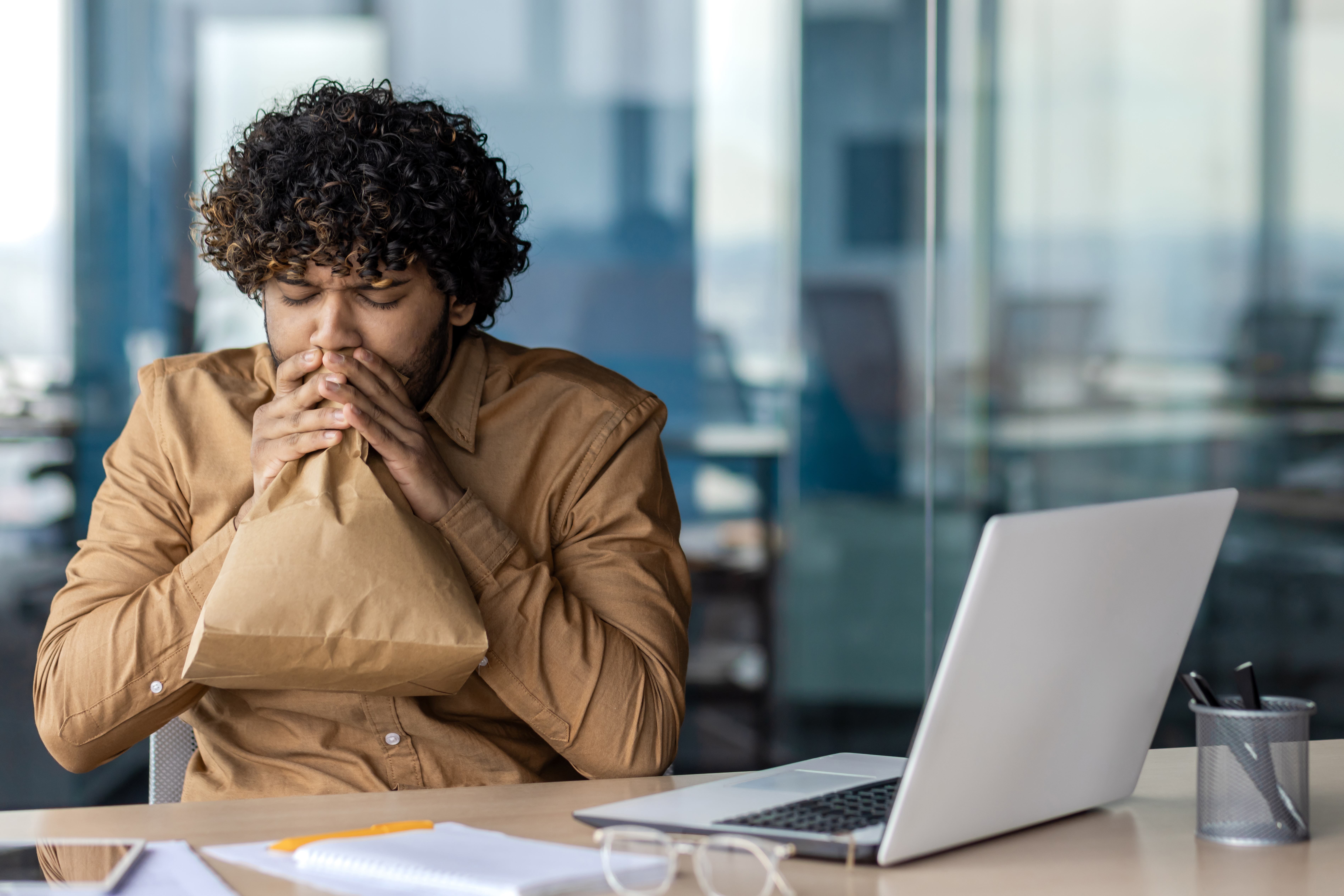 Panic attack in worker inside office at workplace, businessman breathing hard using paper bag, working sitting with laptop