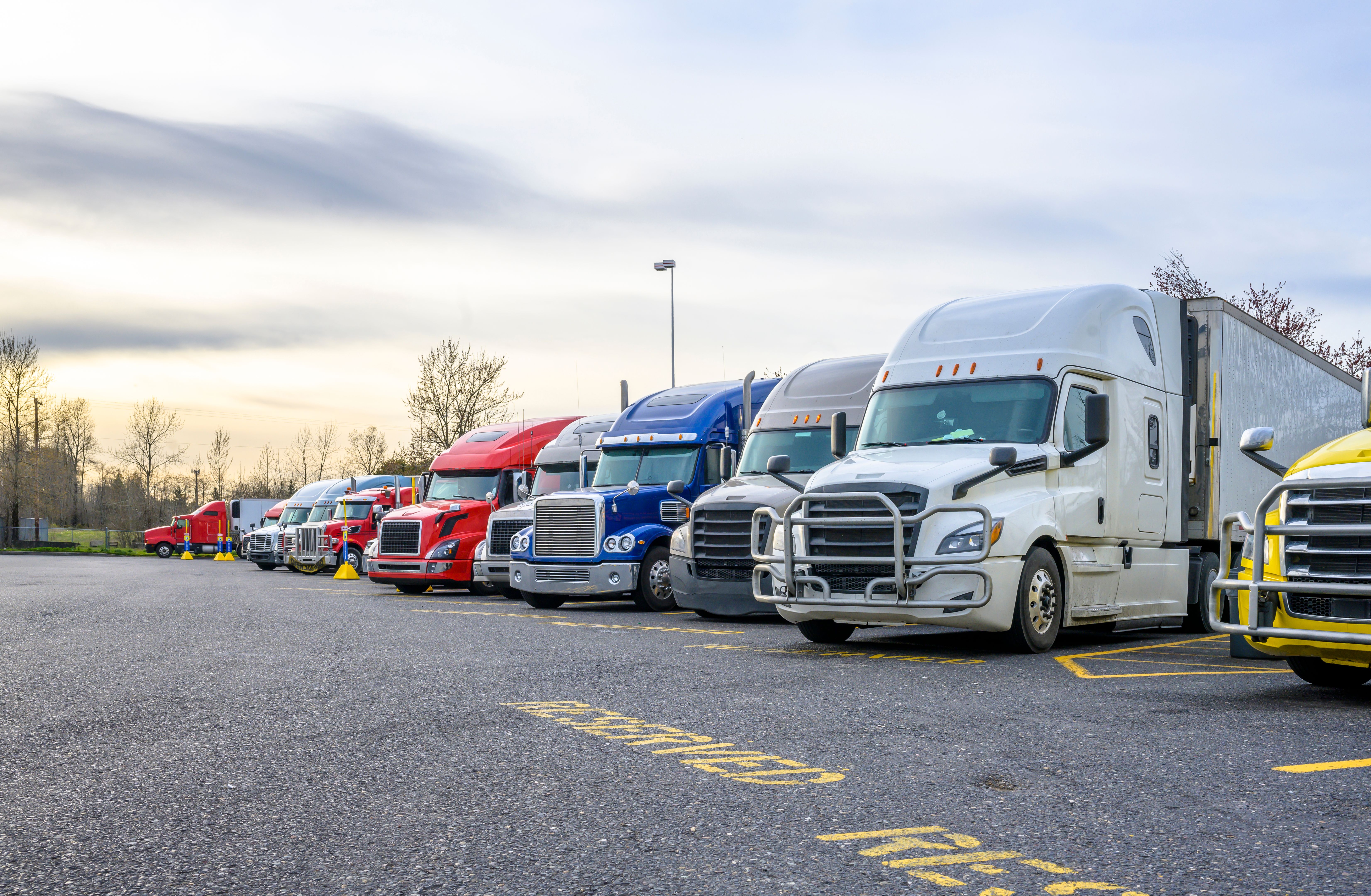 Different big rigs semi trucks with semi trailers standing in row on truck stop parking lot with reserved spots for truck driver rest and compliance with established truck driving regulations