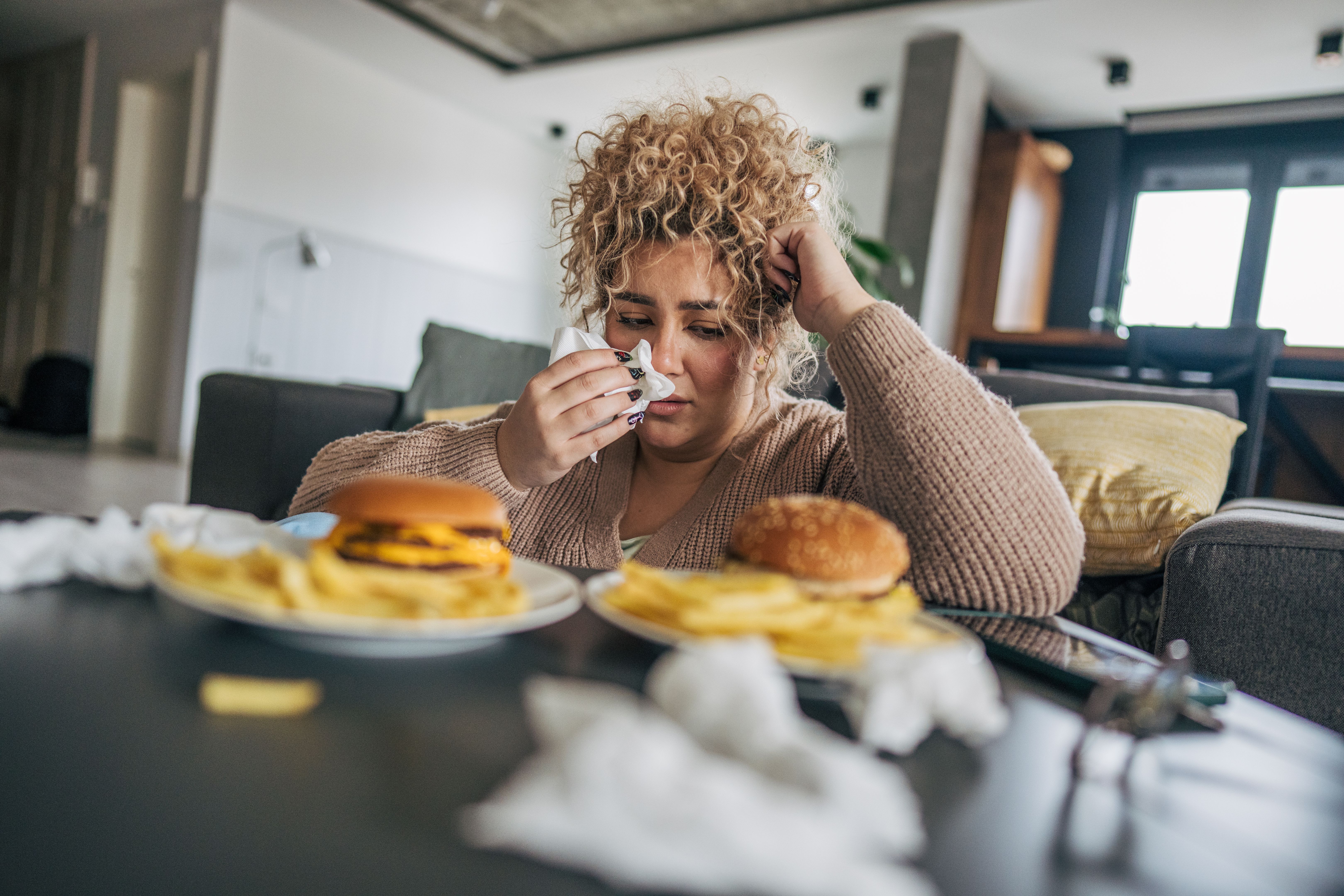 A plus-sized woman with curly hair sitting on the floor, gazing at the two plates of hamburgers and french fries on the table A plus-sized woman with curly hair sitting on the floor, gazing at the two plates of hamburgers and french fries on the table