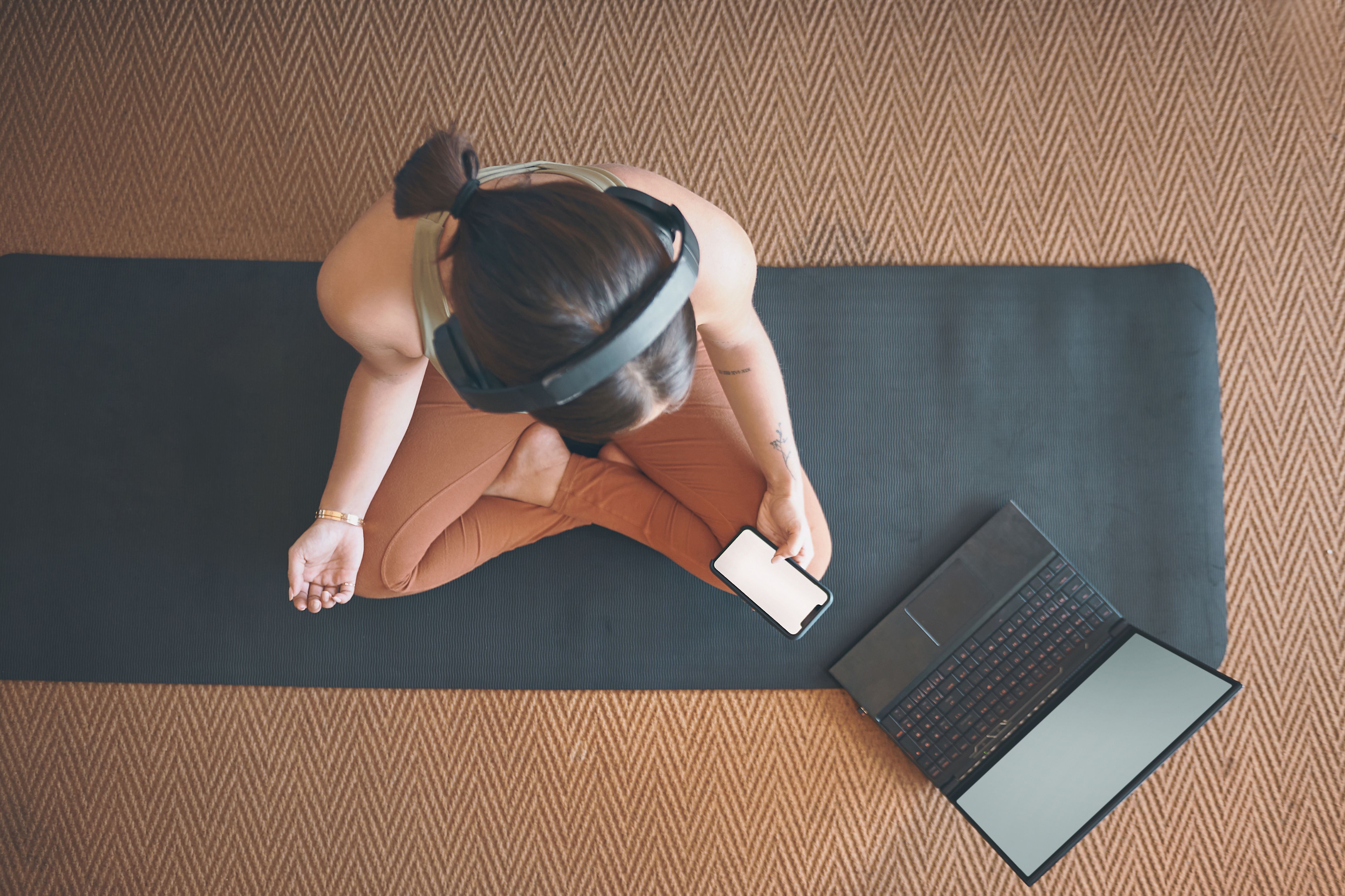 High angle shot of a young woman using a cellphone and laptop while exercising at home