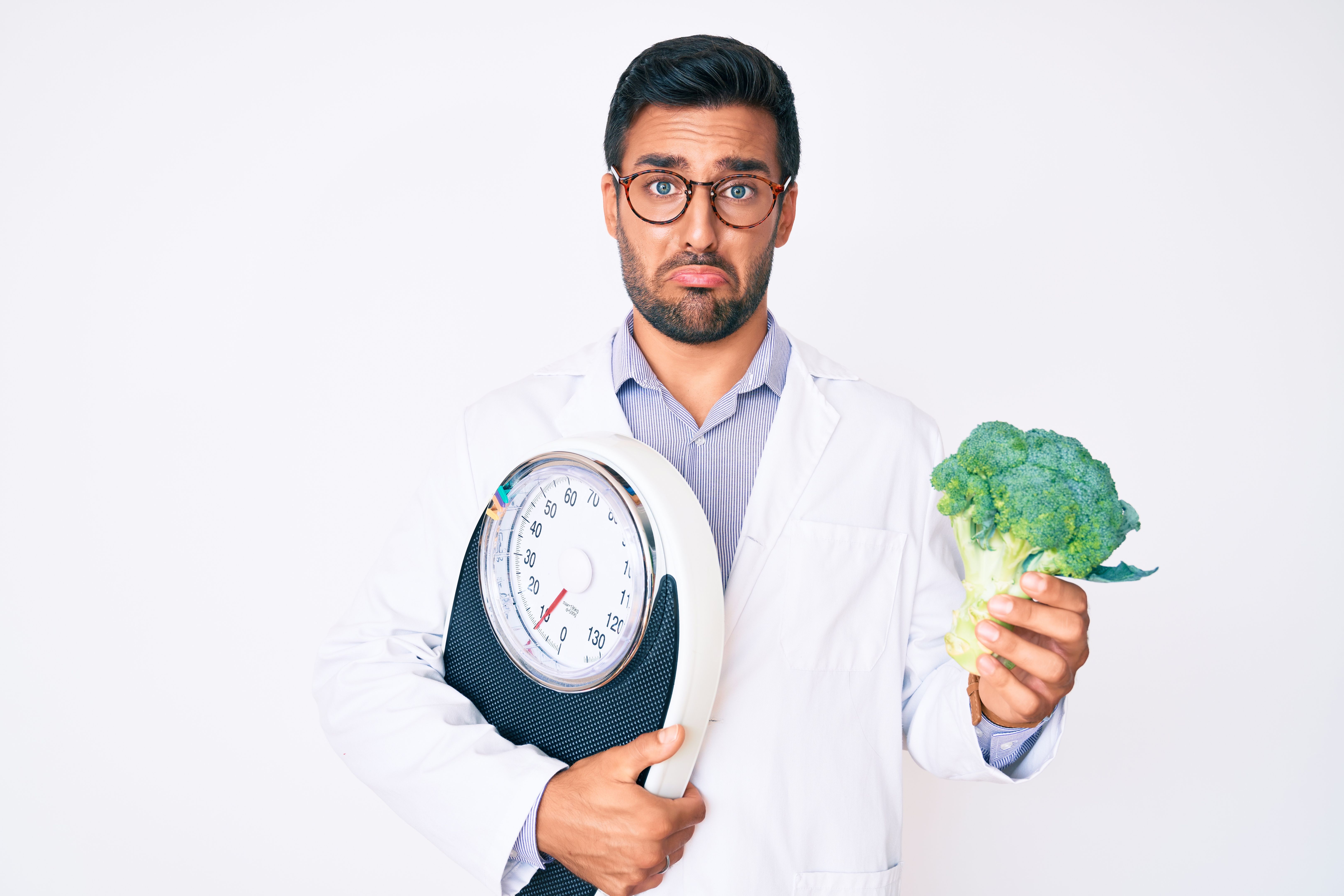 Young hispanic man as nutritionist doctor holding weighing machine and broccoli depressed and worry for distress, crying angry and afraid. sad expression. Young hispanic man as nutritionist doctor holding weighing machine and broccoli depressed and worry for distress, crying angry and afraid. sad expression.
