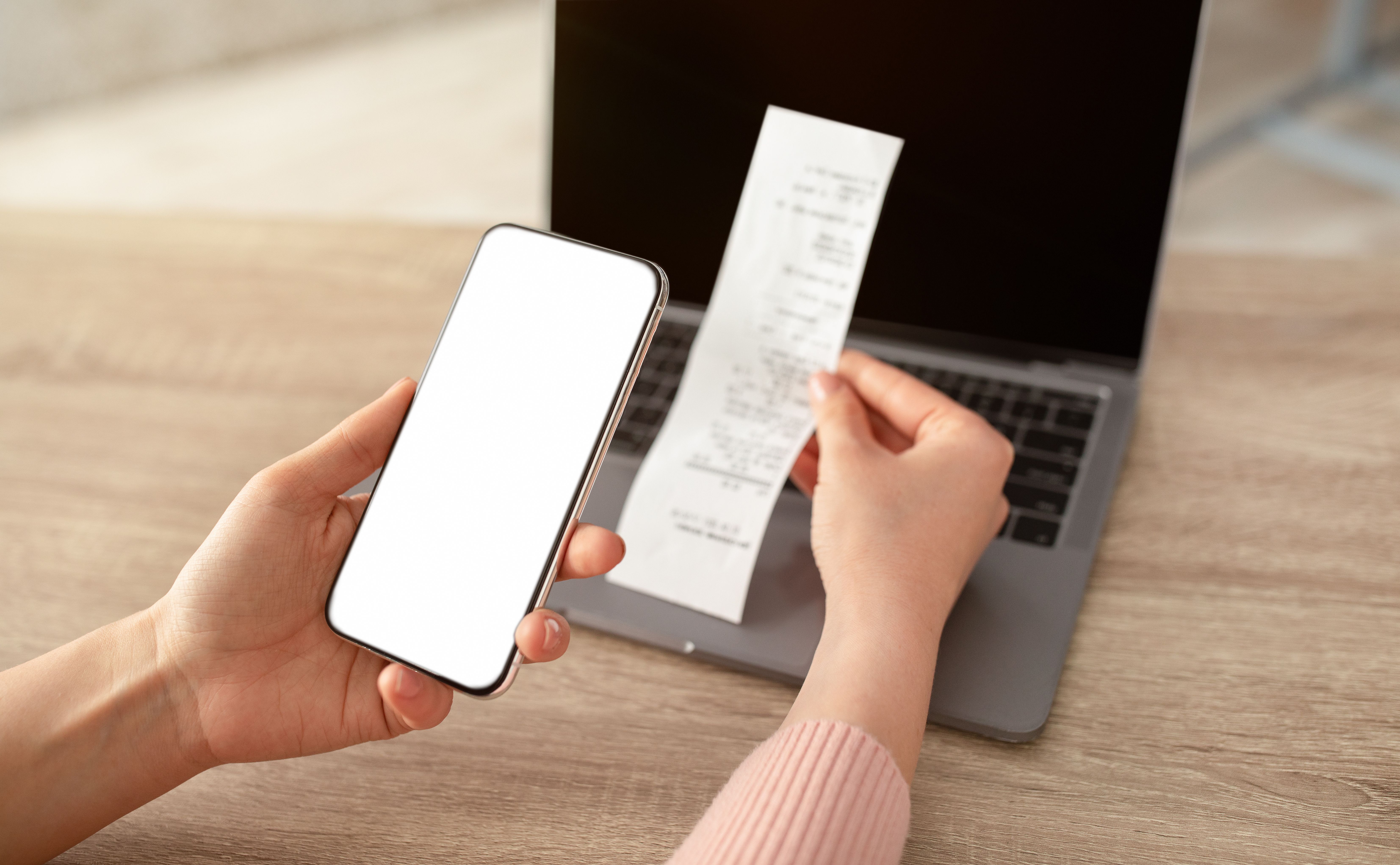 Bill calculating concept. Woman holding smartphone with white blank screen and bill, sitting at workplace with laptop