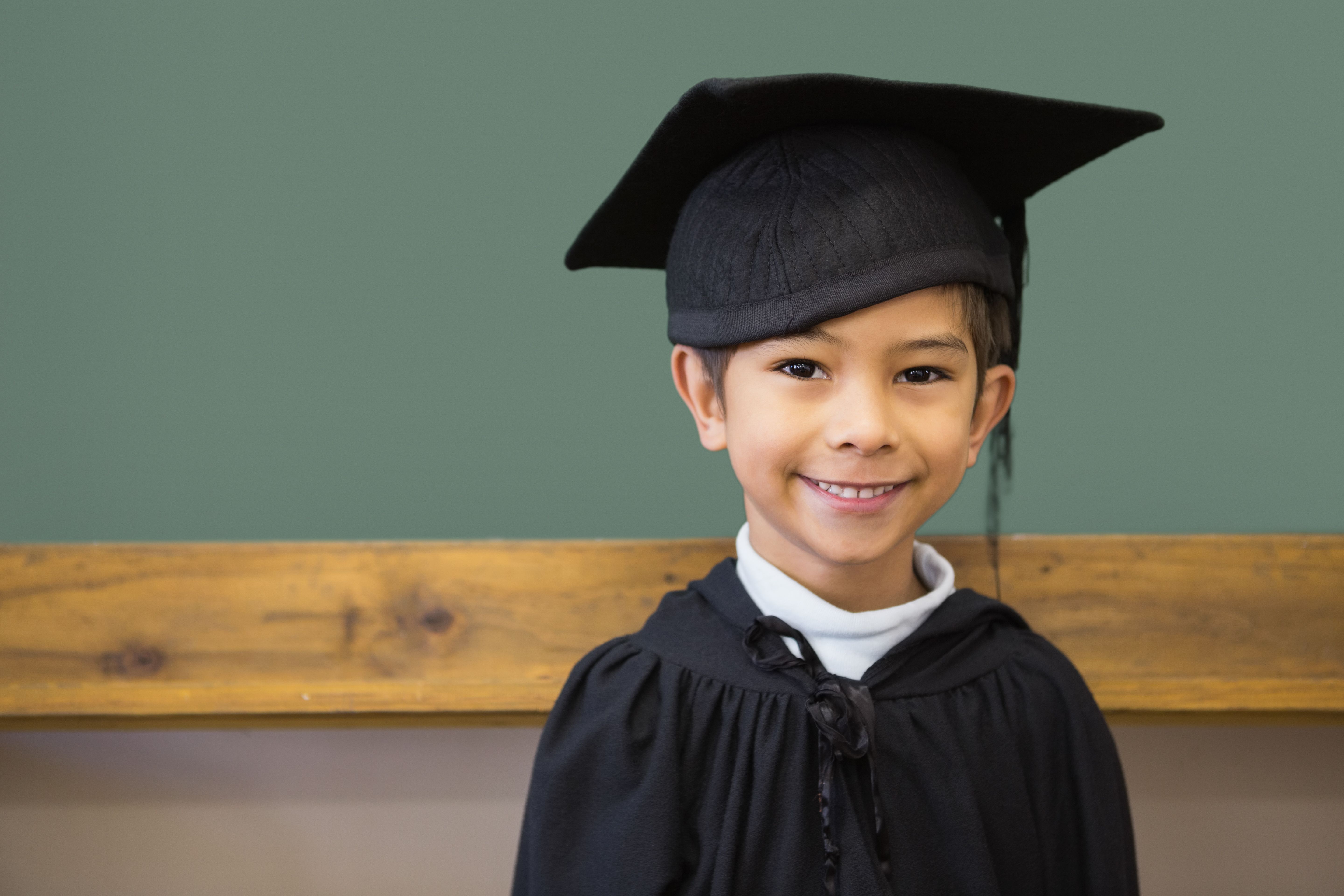 Cute pupil in graduation robe smiling at camera in classroom Cute pupil in graduation robe smiling at camera in classroom