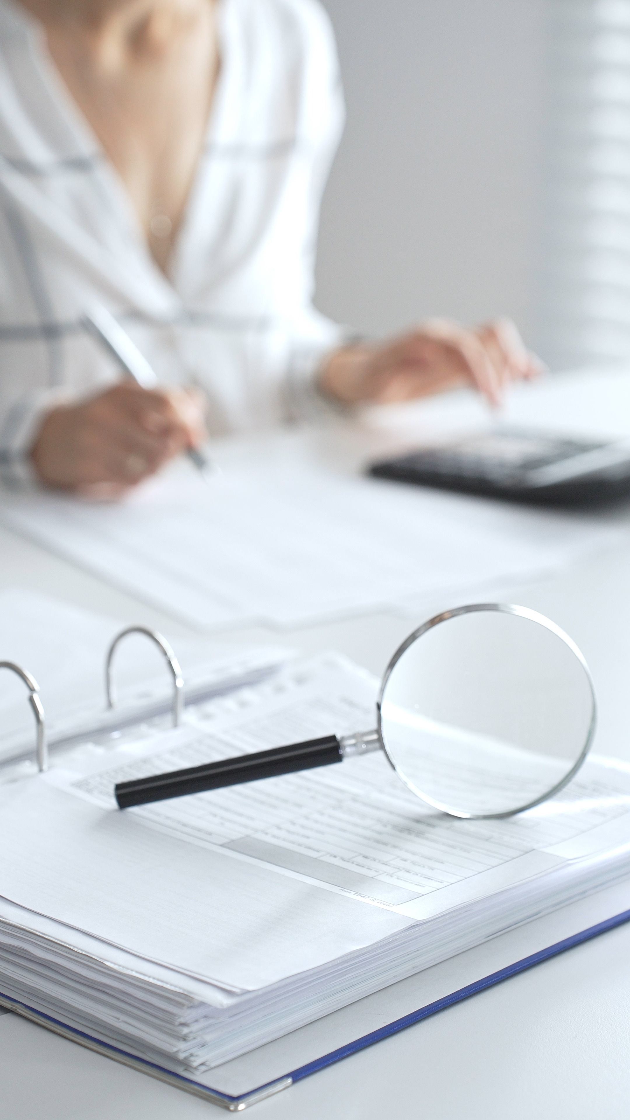 Magnifying glass on financial documents is in focus while an accountant using a calculator and taking notes, emphasizing accounting, finance, close up vertical view. Audit and taxes