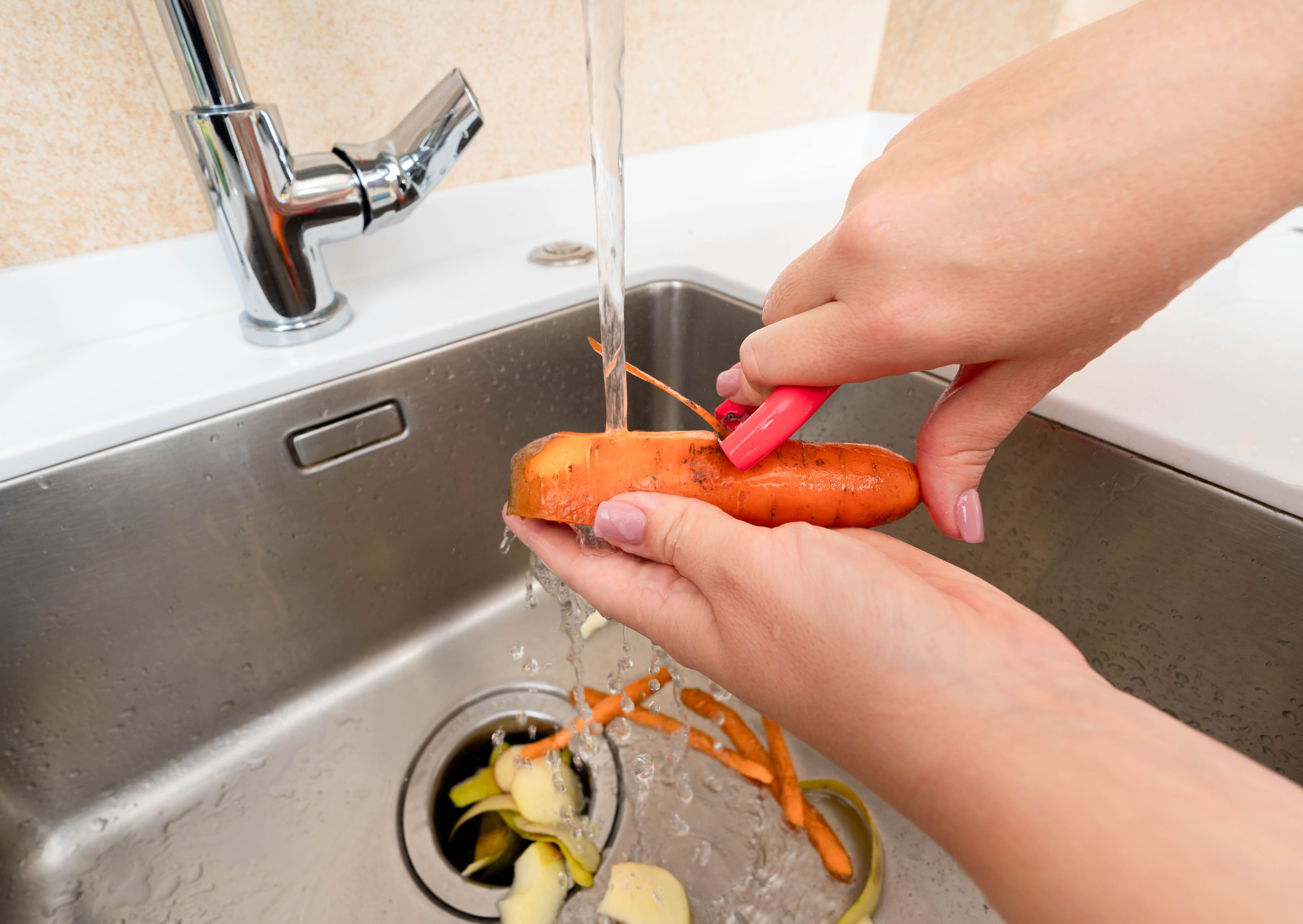 Peeling a fresh carrot under running water with waste collected in a garbage disposer.
