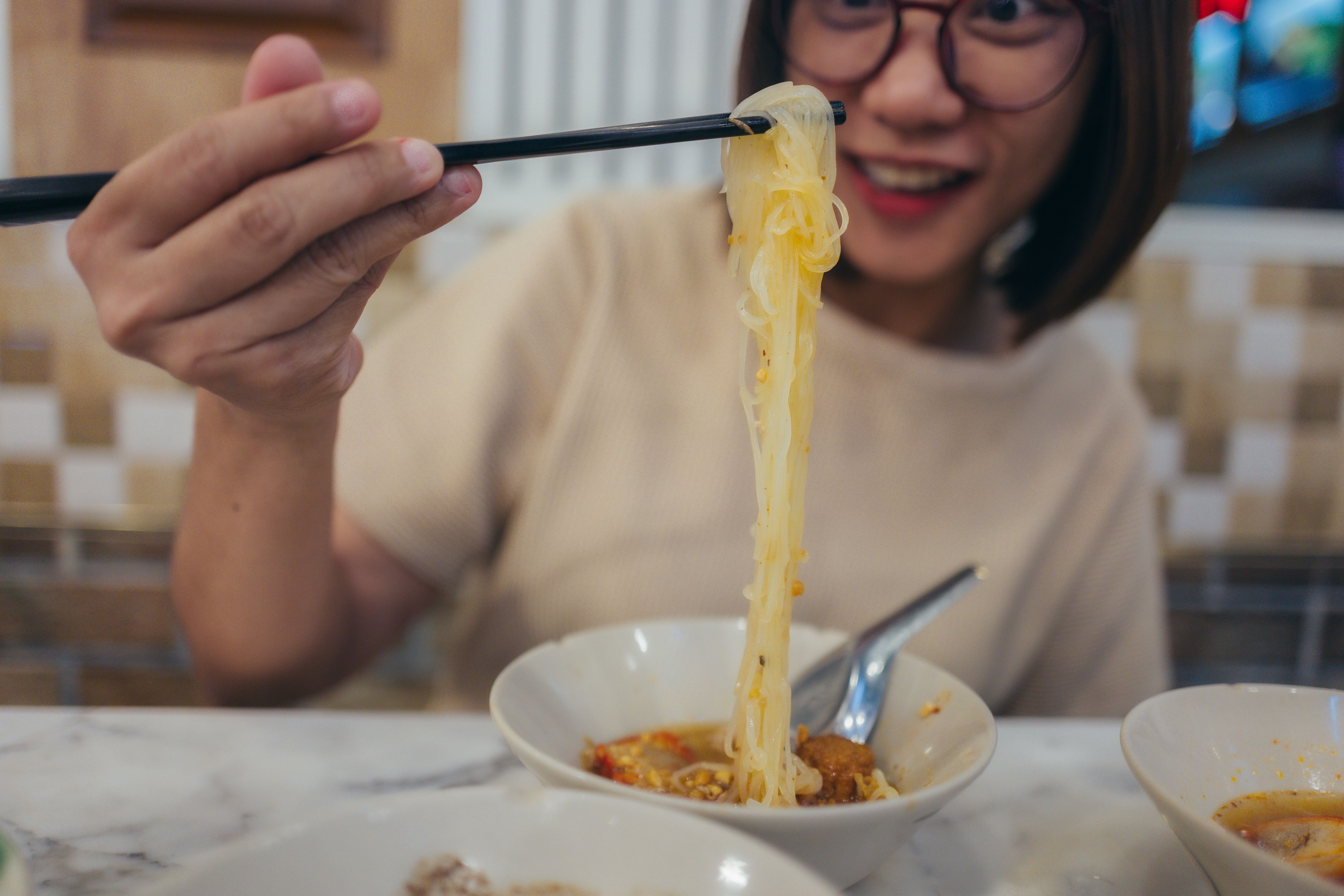 Young woman savoring a delicious bowl of noodles in a restaurant, skillfully lifting them with chopsticks while smiling happily